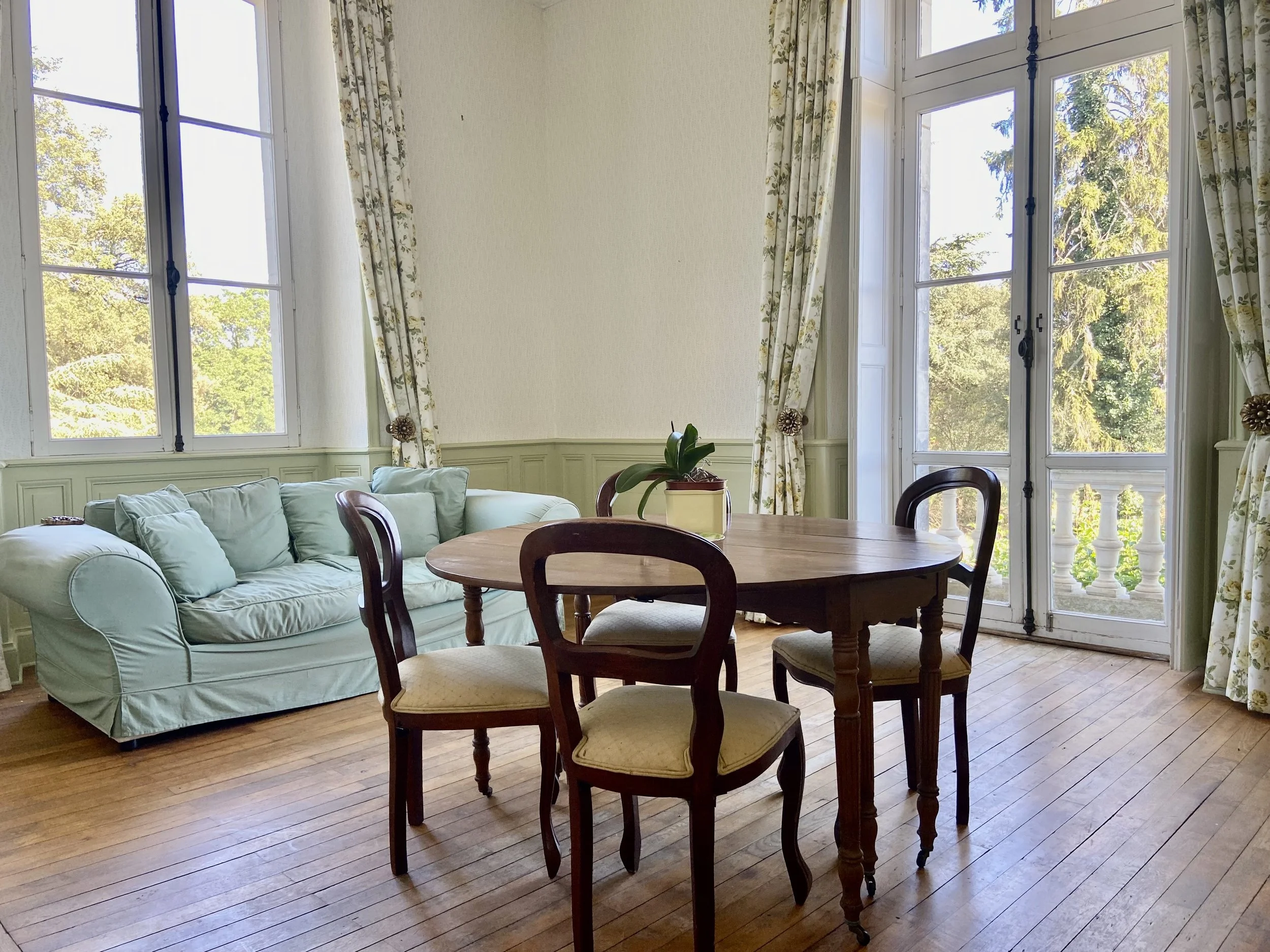 Bright room with a light green sofa, wooden round table, and chairs. Large windows with floral curtains overlook a garden.