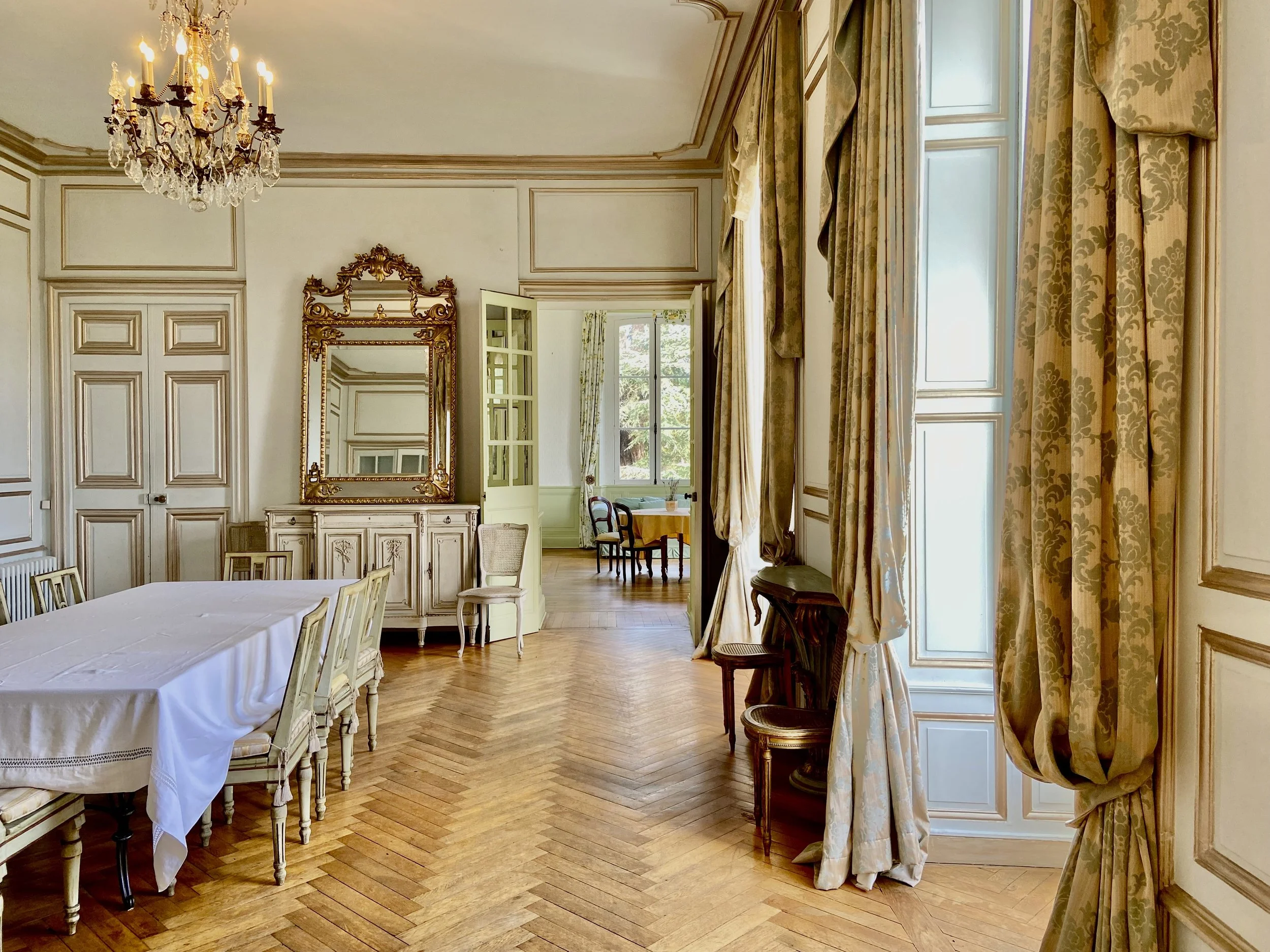 Elegant dining room with vintage furniture, chandelier, large mirror, and long curtains, featuring a table with chairs and parquet flooring.