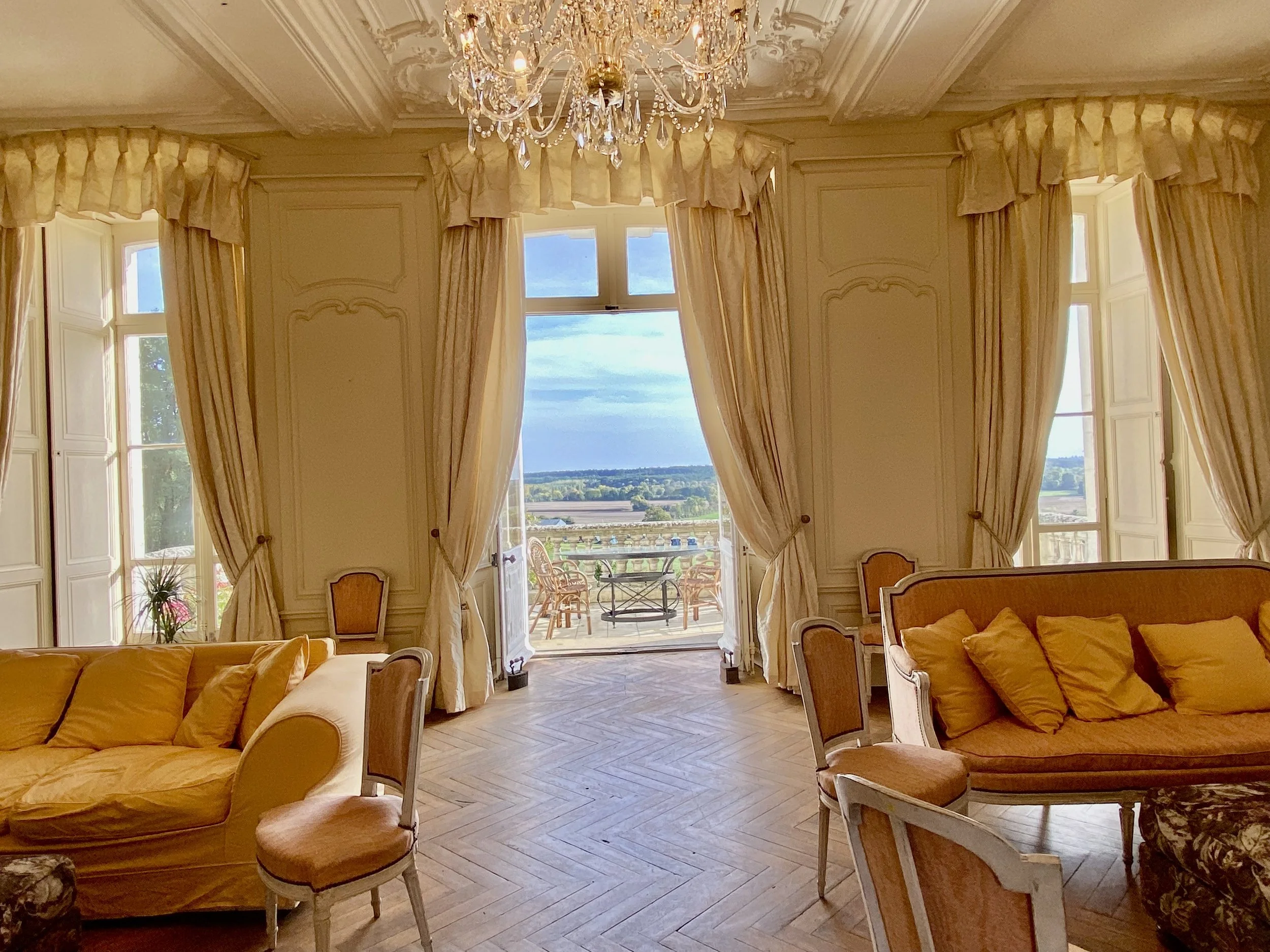 Elegant living room with large windows and cream-colored curtains framing a scenic view. The room features ornate furniture, including sofas and chairs with gold cushions, a parquet floor, and a crystal chandelier.