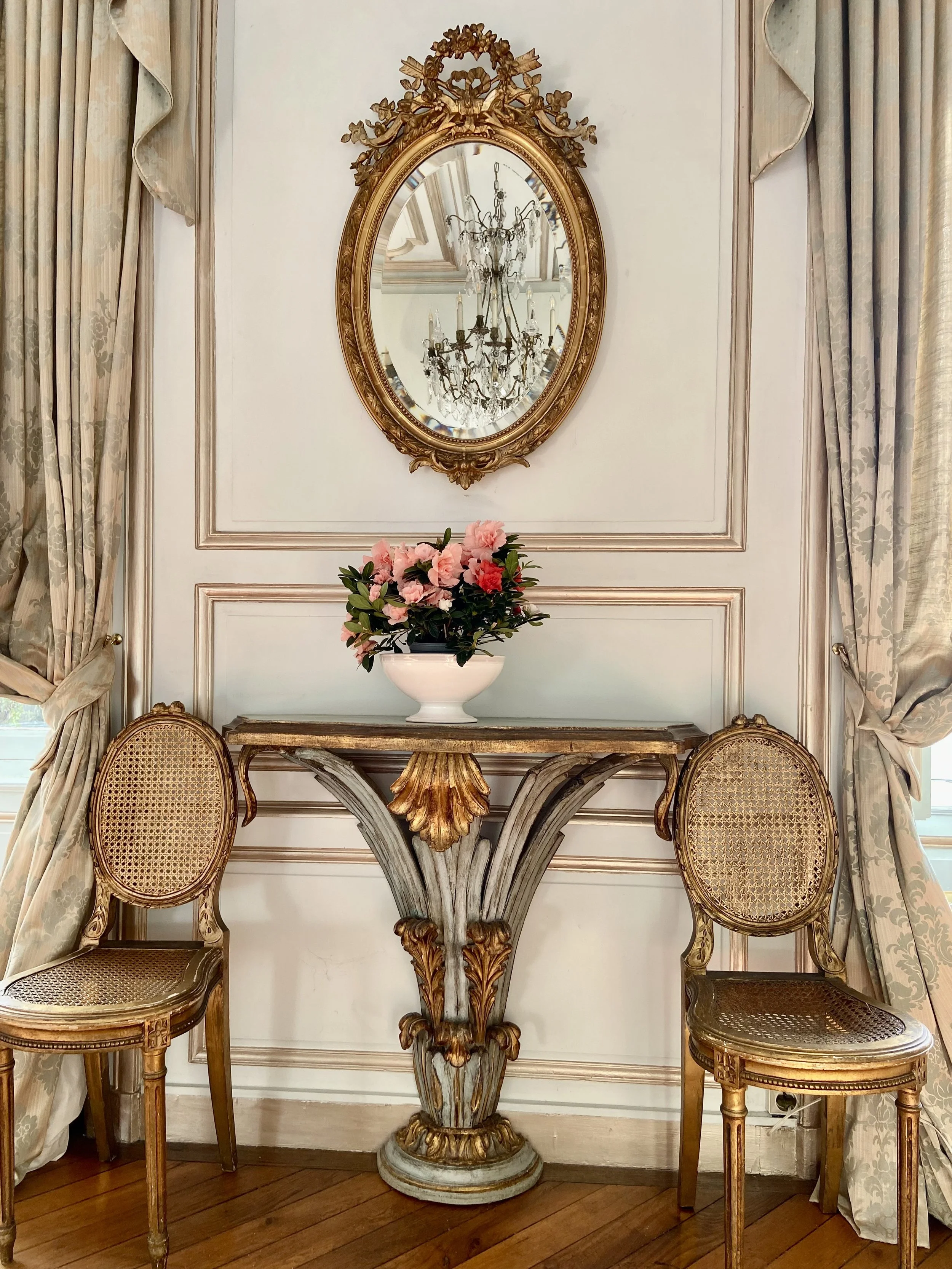 Elegant room with a vintage gold-framed mirror above an ornate console table and floral arrangement, flanked by two upholstered chairs, with elaborate drapery on either side.