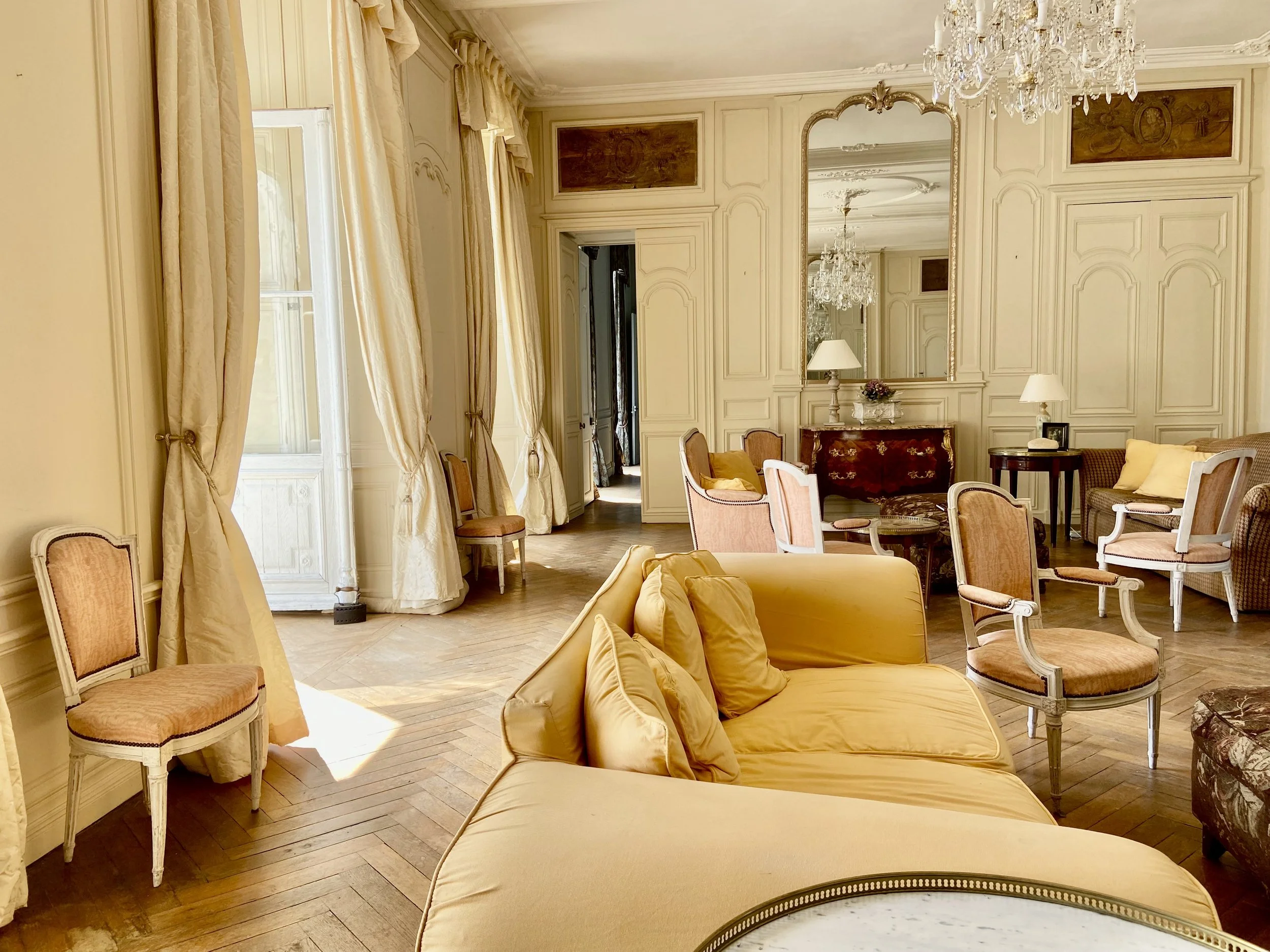 Elegant living room with antique furniture, chandeliers, and large windows with draped curtains.