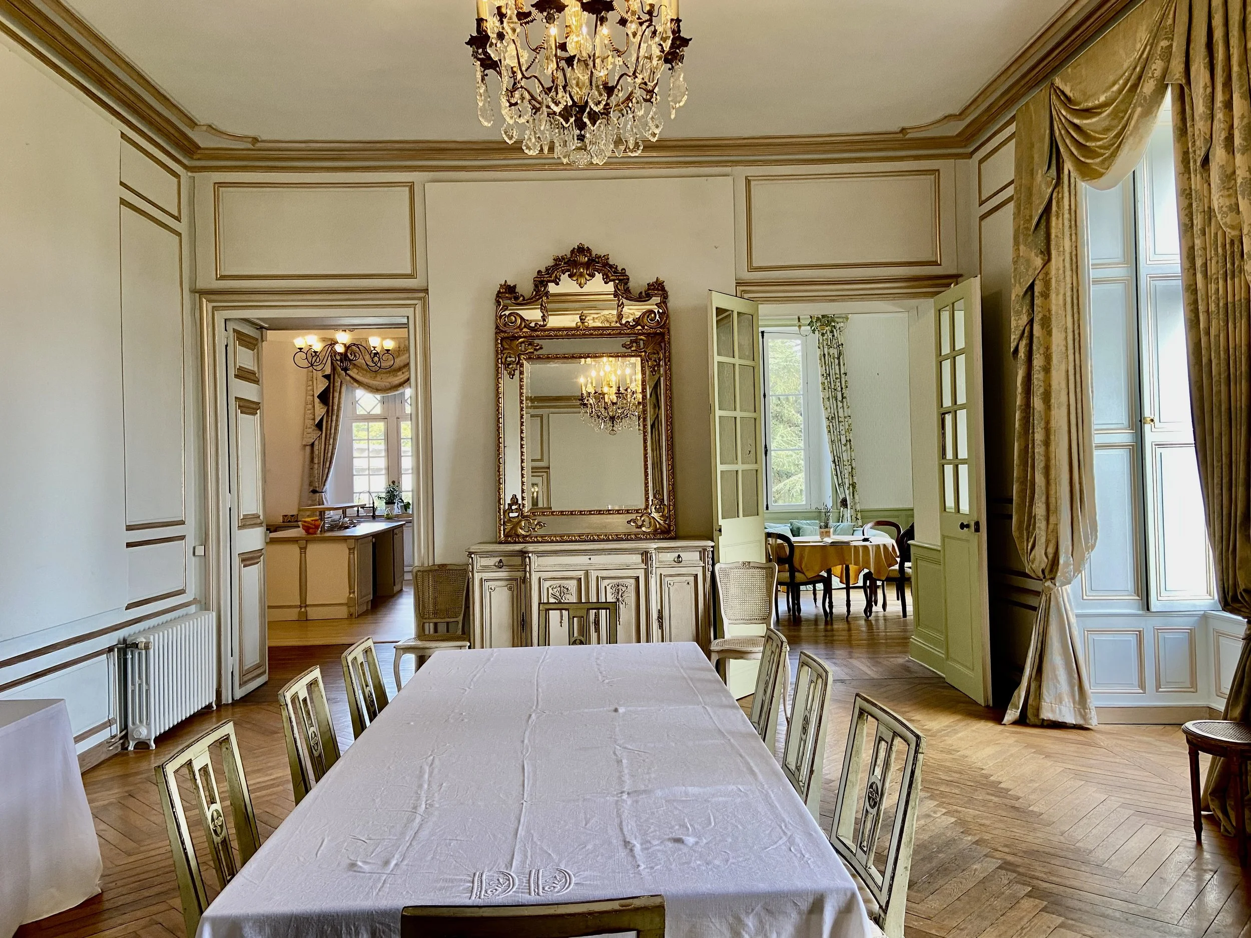 Elegant dining room with white tablecloth, ornate chairs, a large mirror, chandelier, and draped curtains.