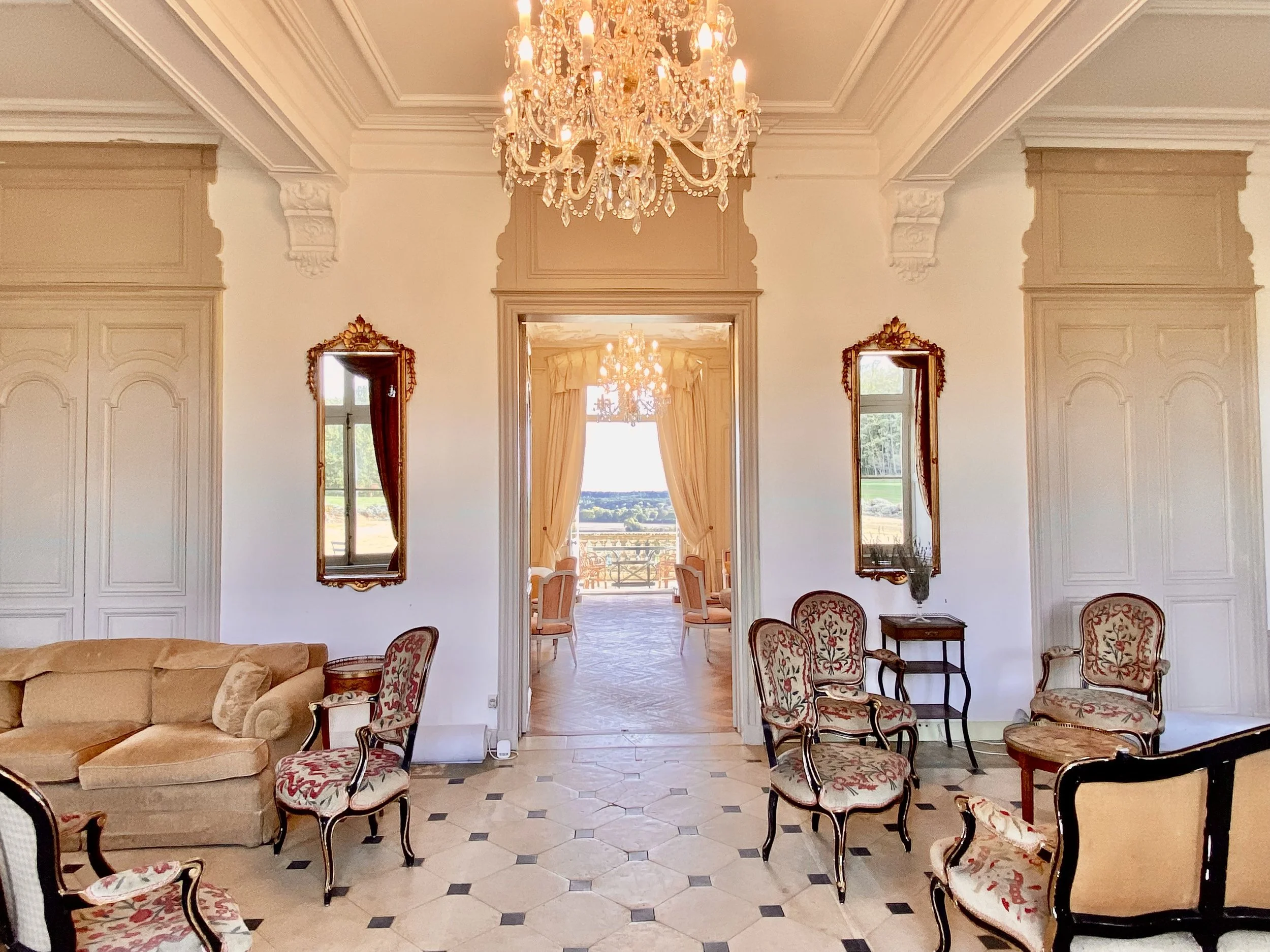 Elegant room with vintage furniture and a chandelier, featuring ornate armchairs, a plush sofa, decorative mirrors, and tall windows overlooking a scenic view.