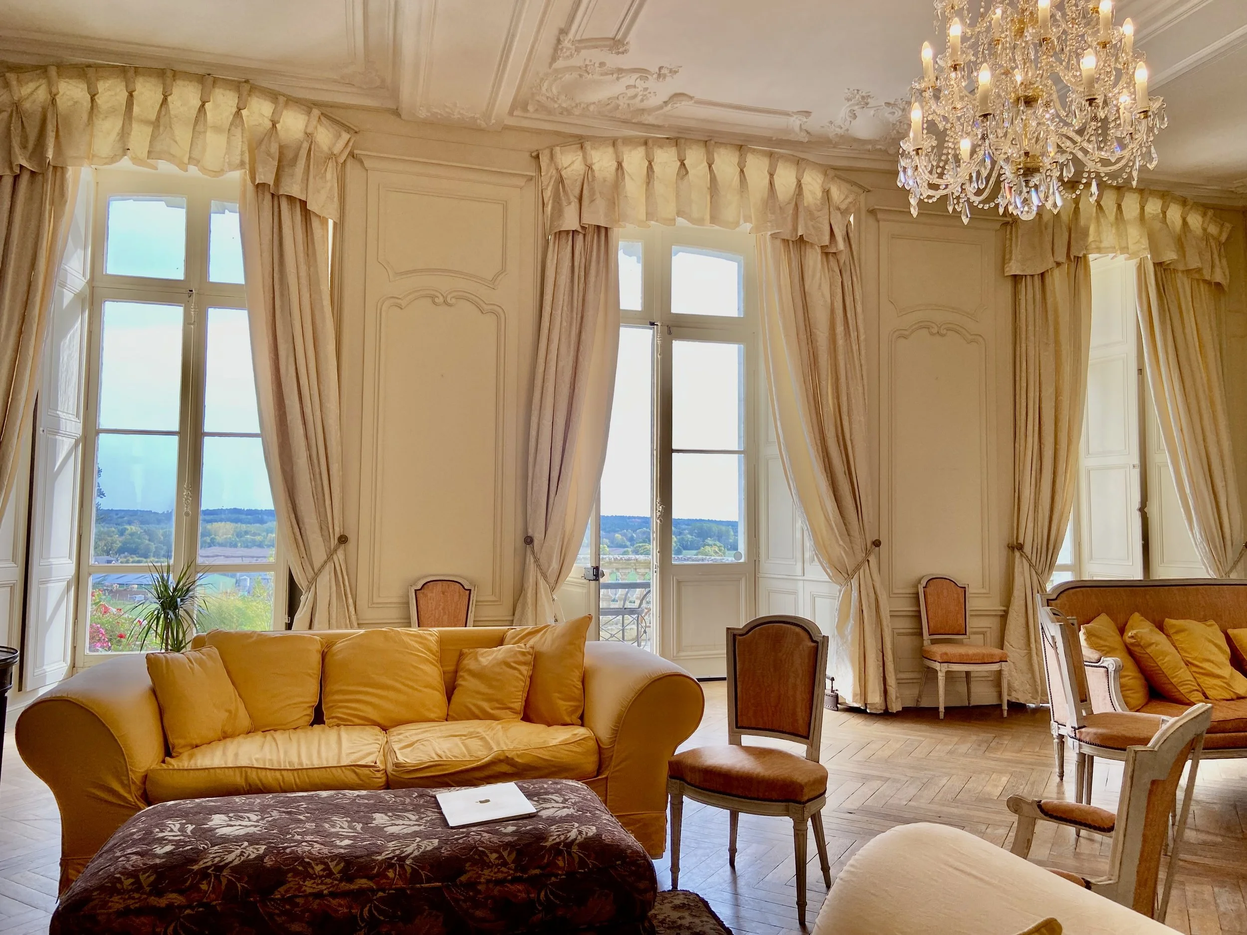 Elegant living room with large windows, cream curtains, classic sofas, chairs, and a crystal chandelier.