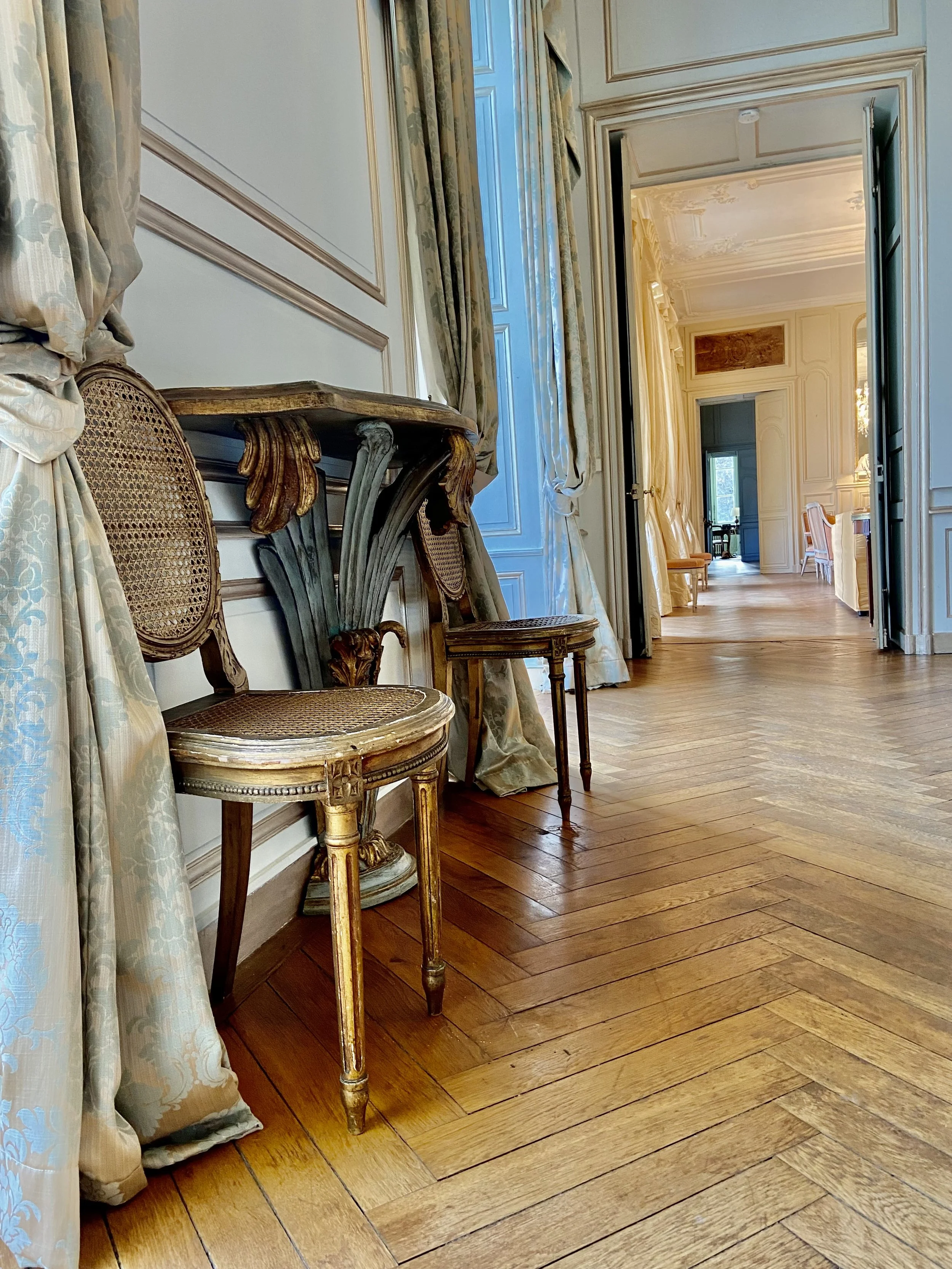 Elegant interior of a historic room featuring ornate chairs, a decorated console table, wooden parquet flooring, and luxurious drapes.