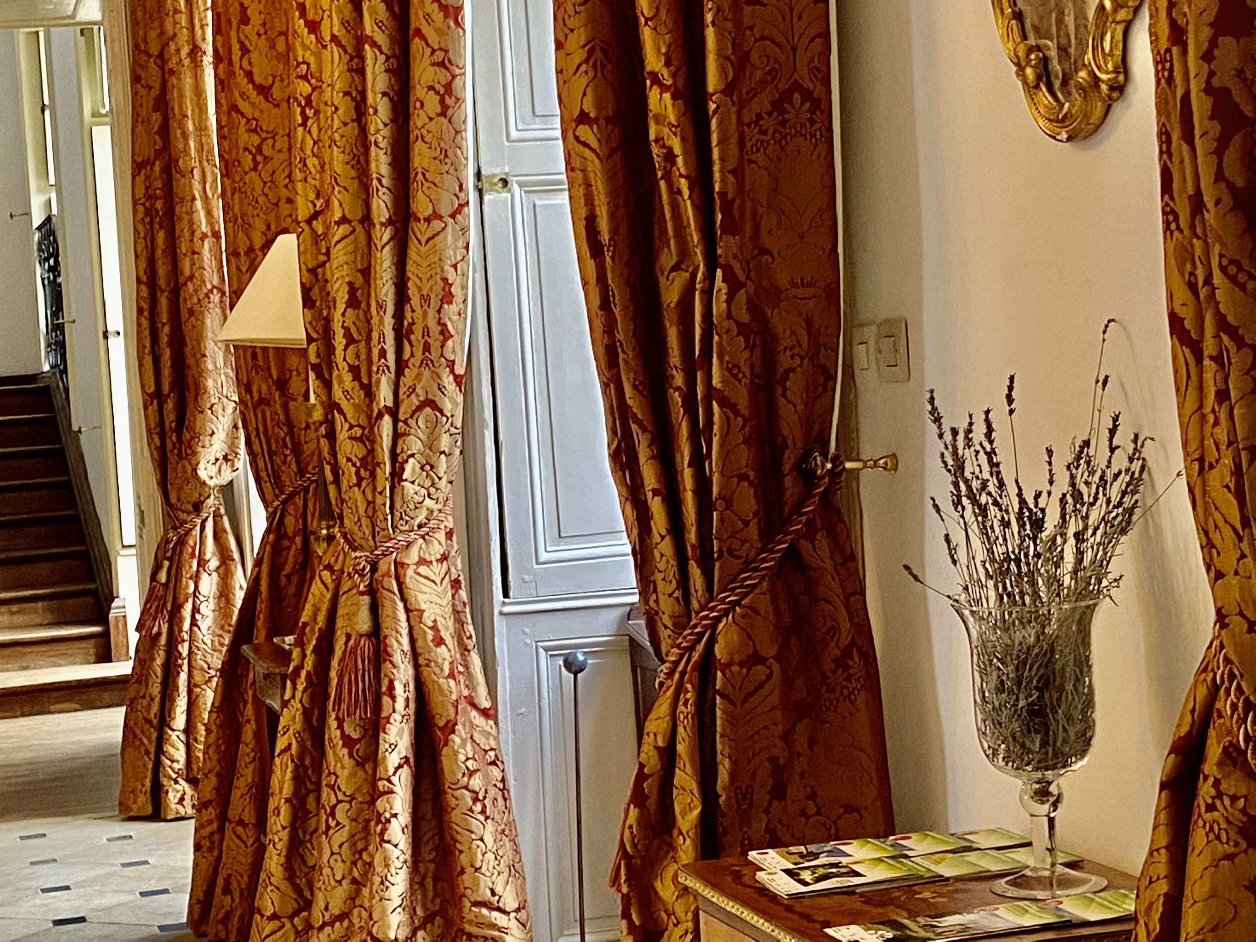 Elegant interior with ornate, gold-patterned curtains, a marble-topped table with lavender flowers in a glass vase, and a staircase in the background.