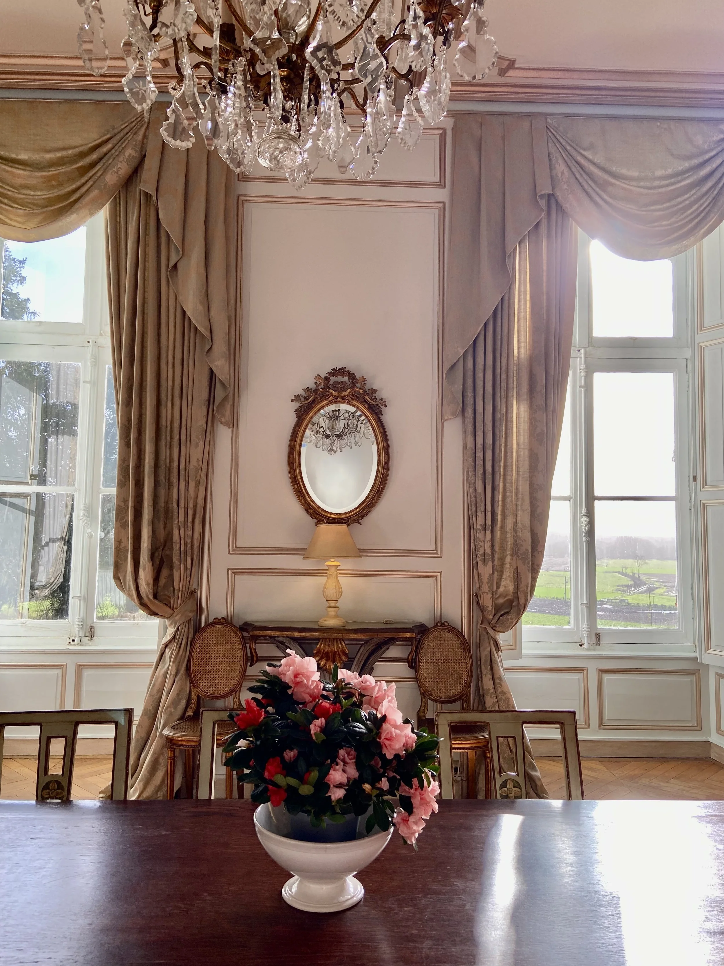 Elegant dining room with a chandelier, wall mirror, large windows, and a table centerpiece of pink flowers in a white vase.