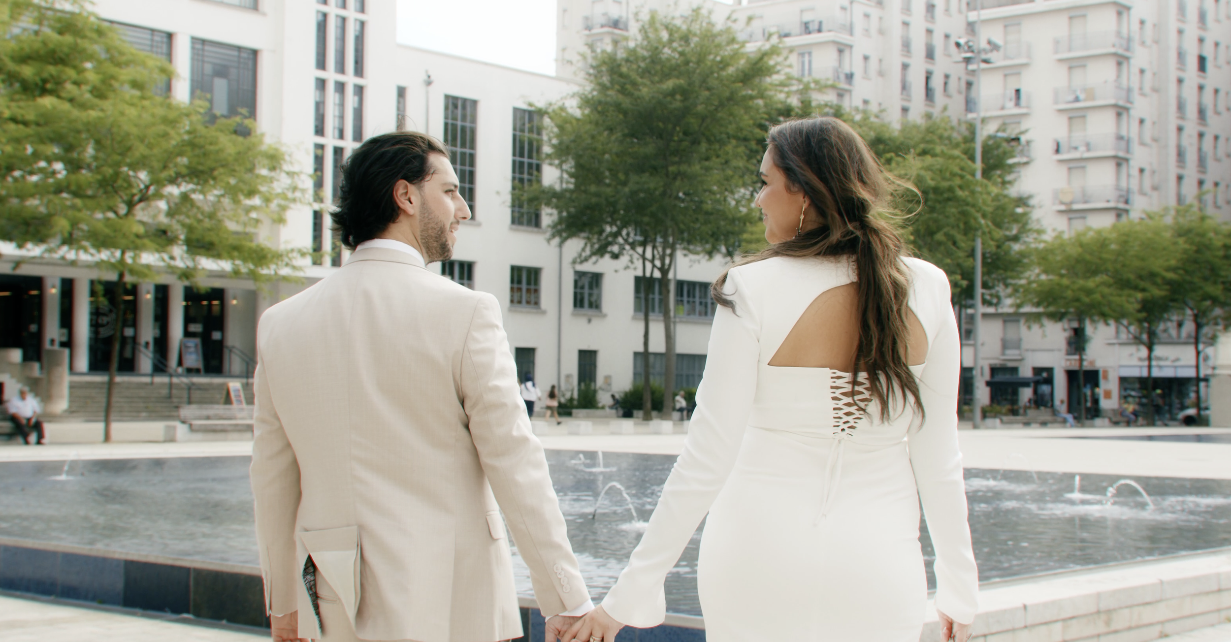 A couple holding hands and walking in an outdoor urban area with fountains and modern buildings, wearing formal attire.