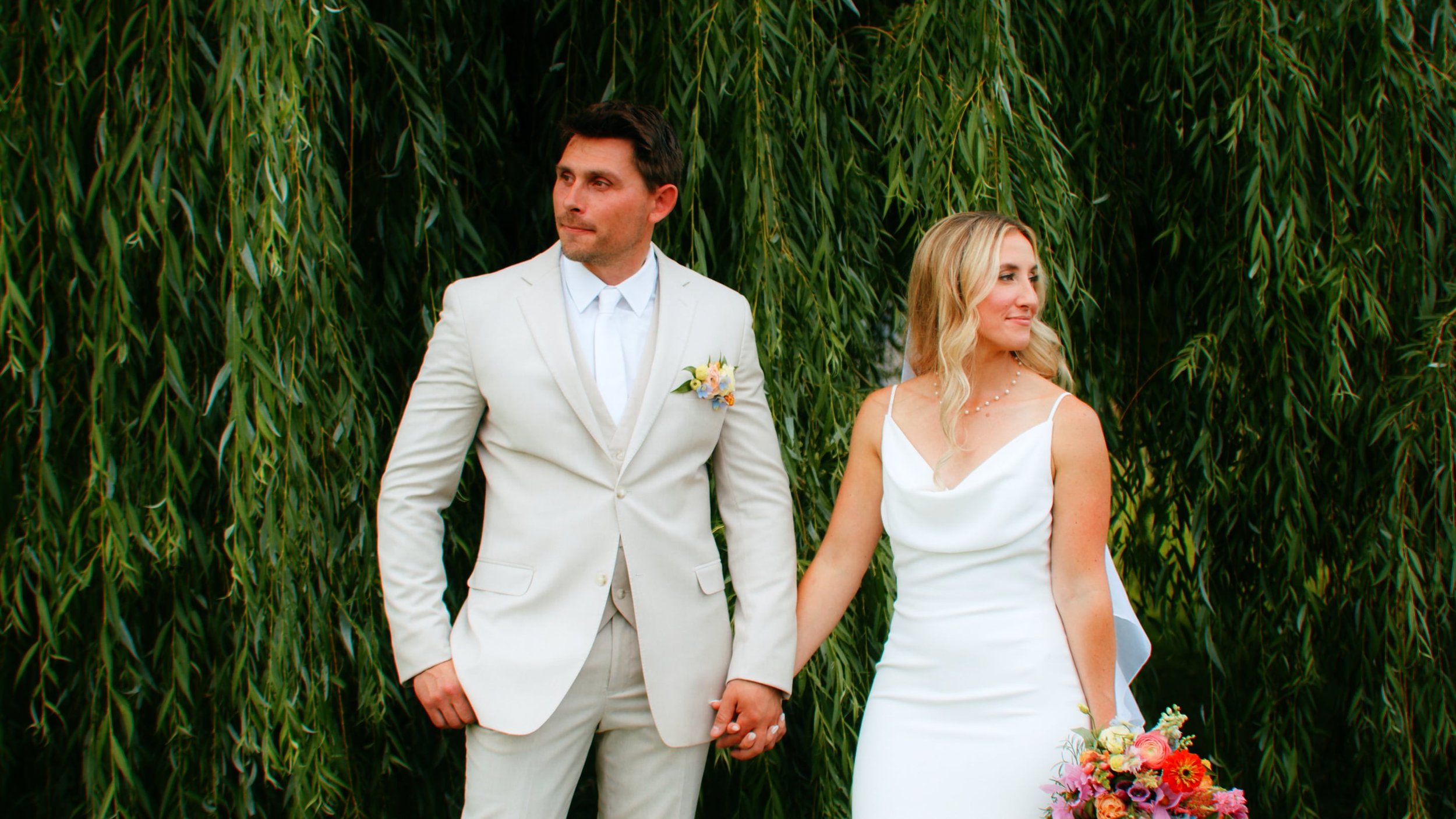 A bride and groom holding hands, standing outdoors in front of green foliage, with the groom in a light cream suit and the bride in a white dress holding a colorful bouquet.