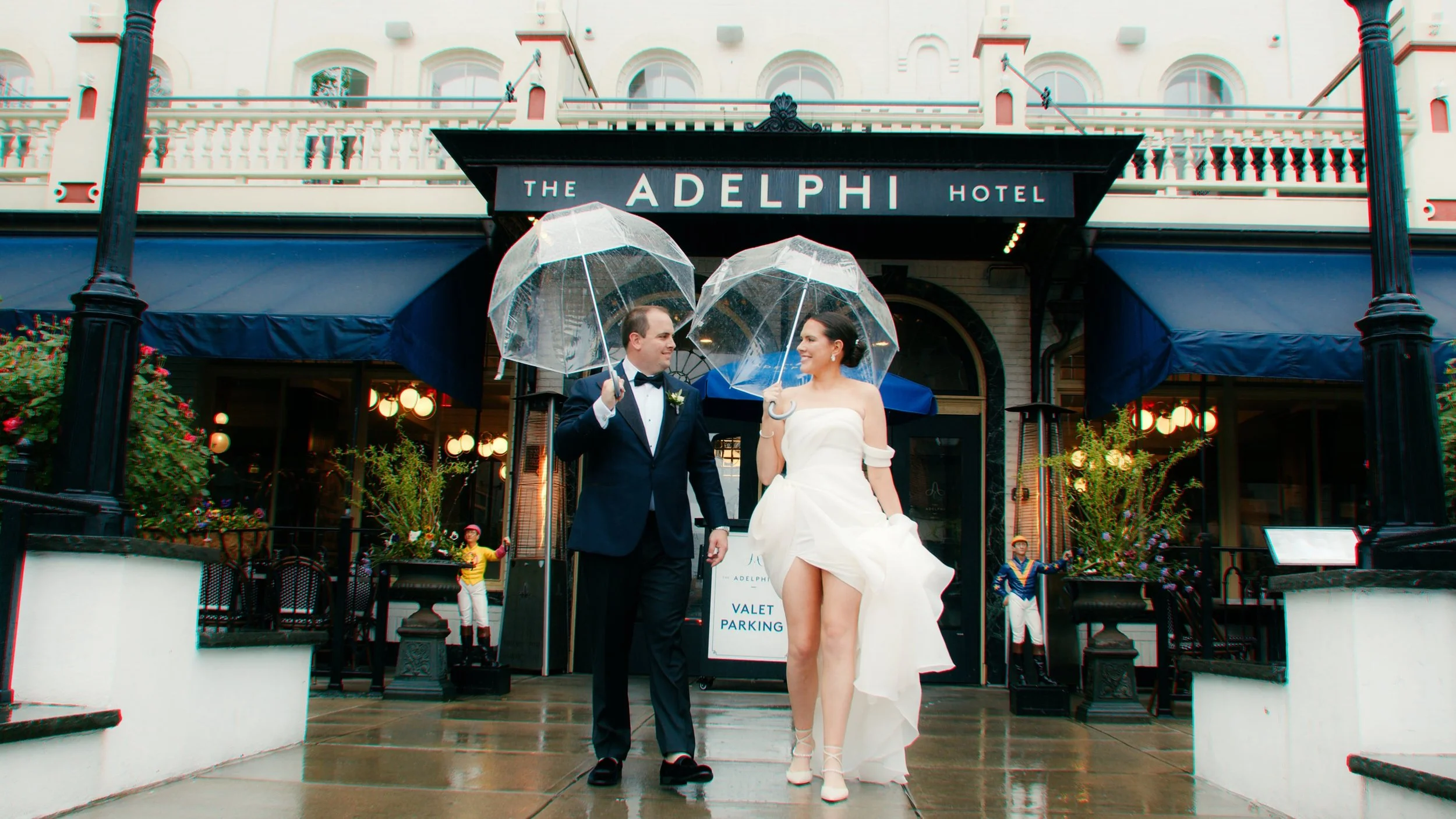 A bride and groom dressed in wedding attire standing outside The Adelphi Hotel, holding transparent umbrellas and smiling at each other on a rainy day.
