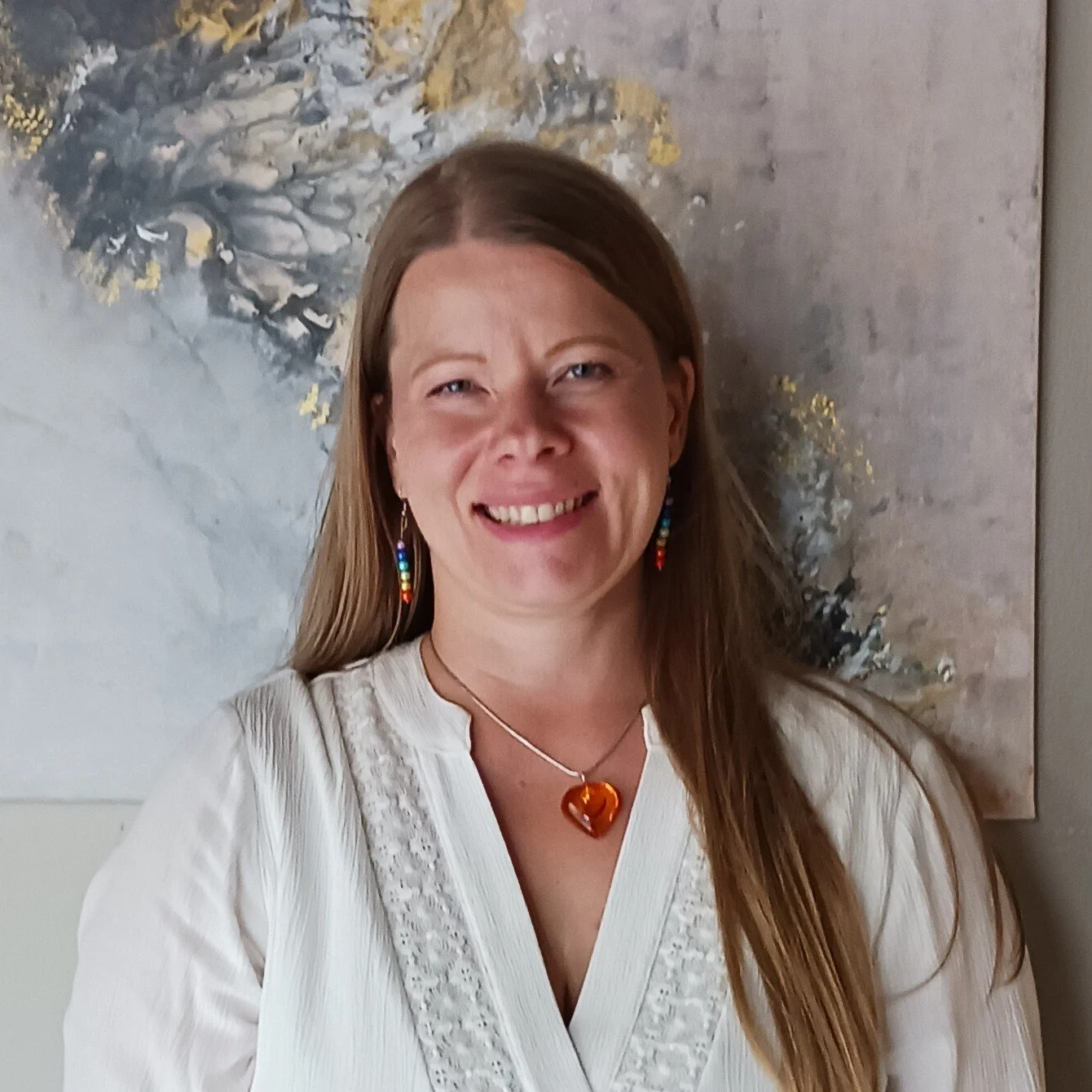 Hymyilevä nainen valkoisessa paidassa, värikkäillä korviksilla ja sydänriipuksella, taustalla abstrakti seinämaalaus.