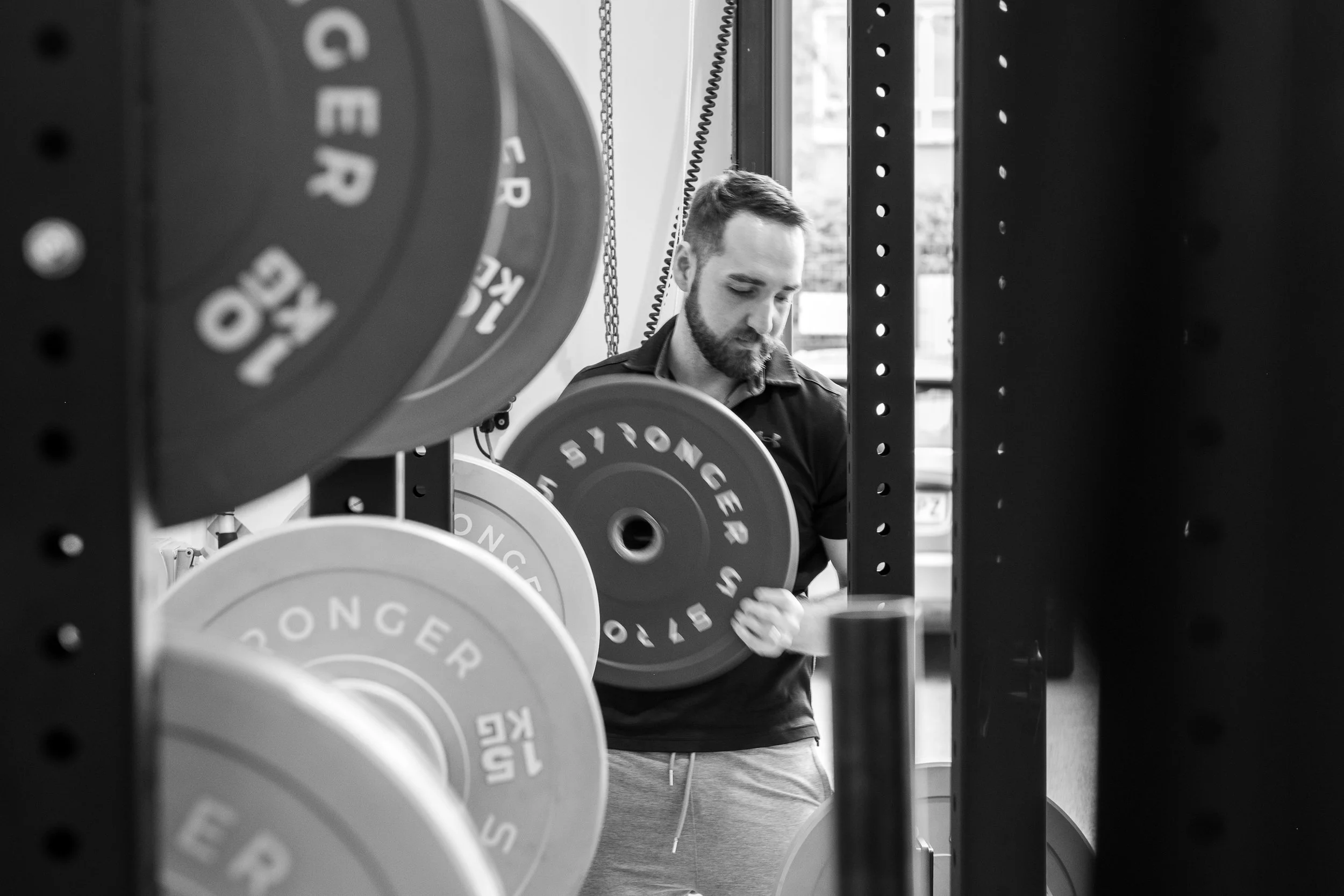 A man with a beard and short hair in workout attire standing among weightlifting bumper plates on a squat rack, in a gym.