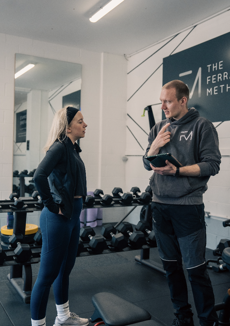 A woman and a trainer having a discussion in a gym, with dumbbells and exercise equipment in the background.