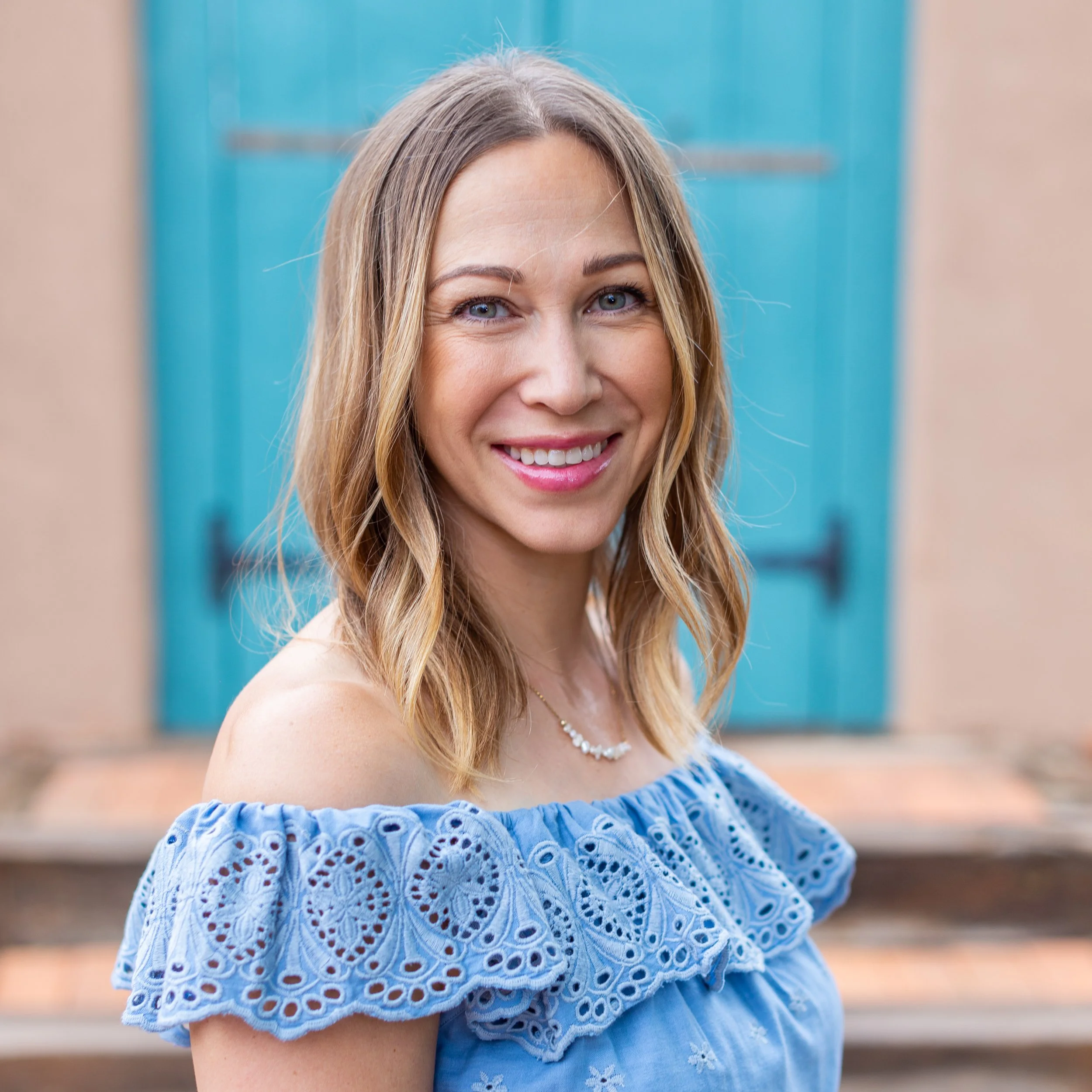 A woman with blonde hair and blue eyes wearing a blue eyelet dress and a pearl necklace, smiling outdoors with a building and steps in the background.
