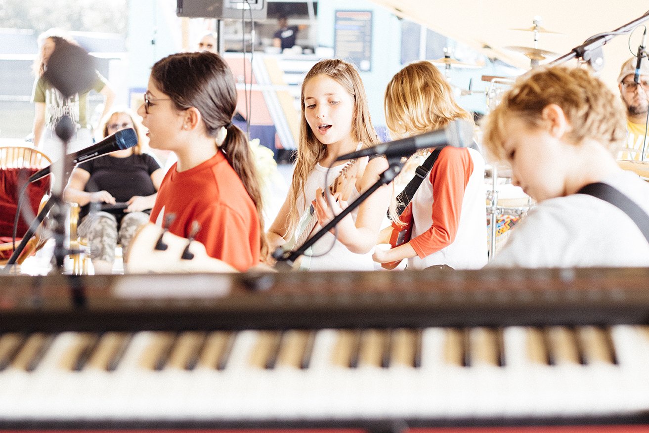 Children performing music on stage, playing guitars and singing, with musical instruments and microphones, at an indoor event.