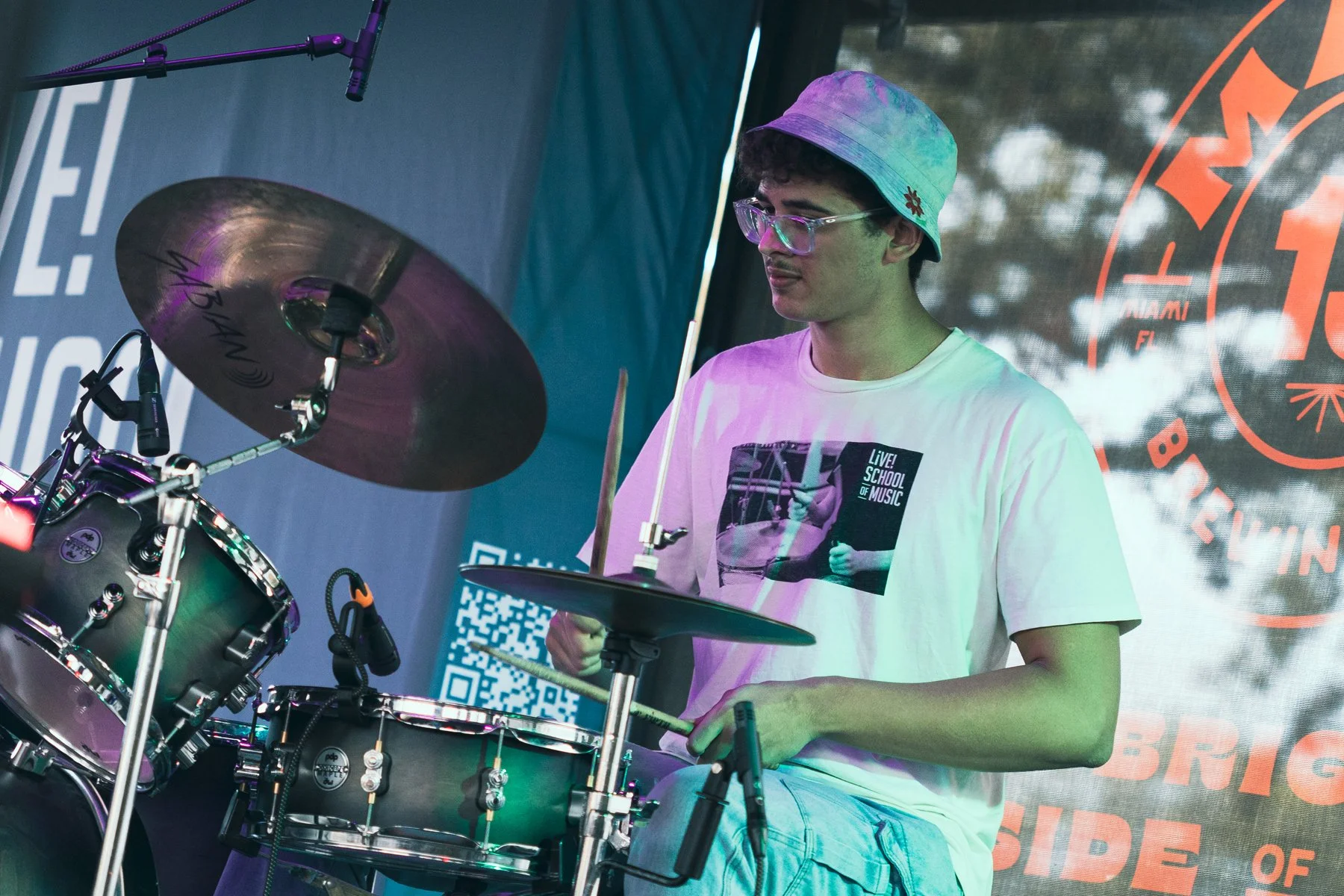 A young man wearing glasses, a tie-dye bucket hat, and a white T-shirt with a graphic that reads 'Live! School of Music' is playing drums during a performance on a stage with a blue backdrop and a QR code.