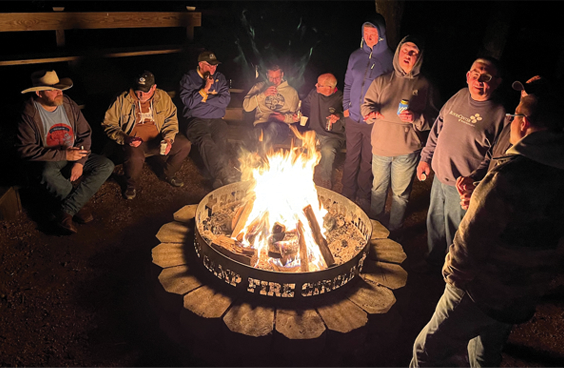 A group of people gathered around a campfire at night, some standing and some sitting, in an outdoor setting.