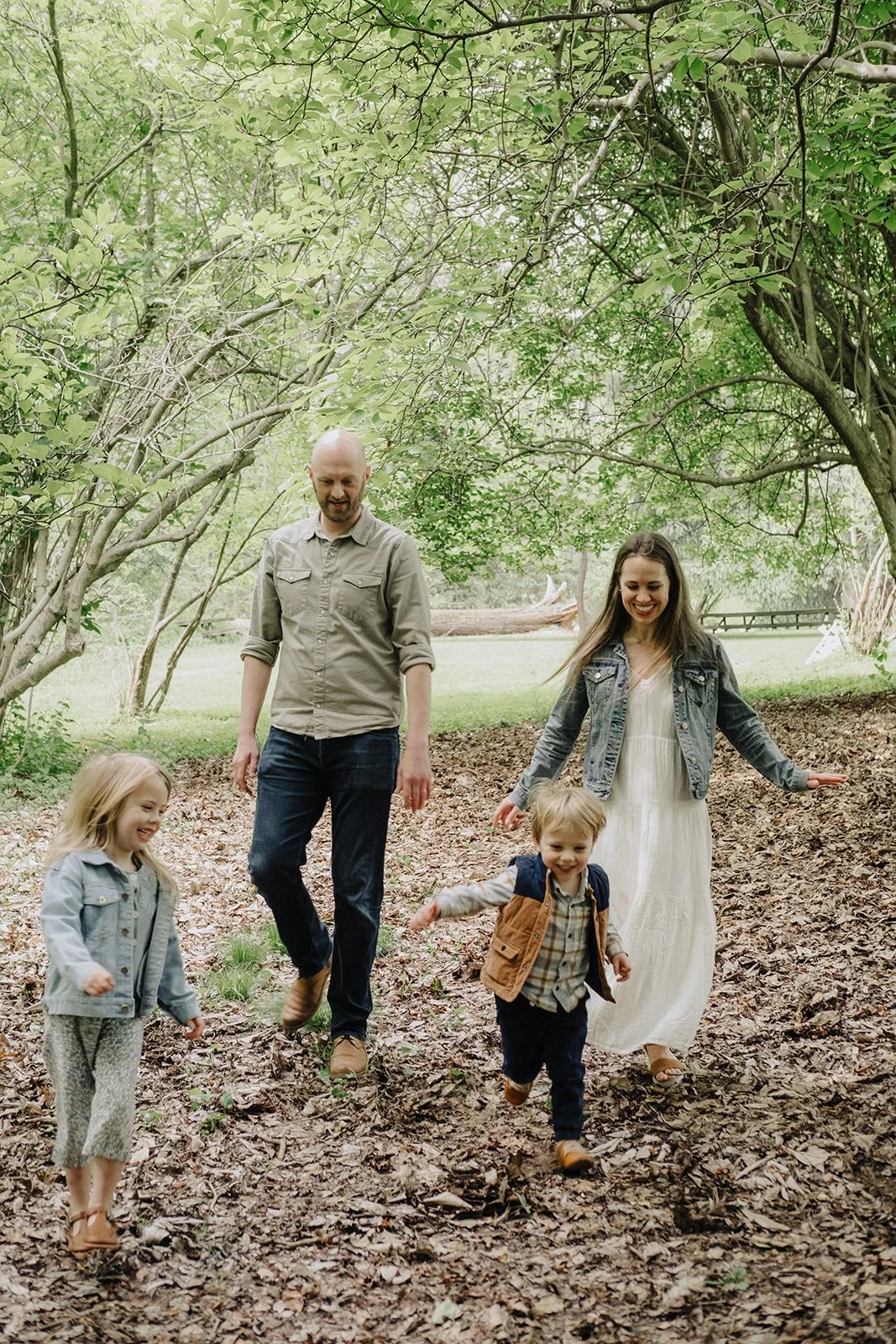 A family of four with two children, walking and playing outdoors on a leaf-covered trail under lush green trees.