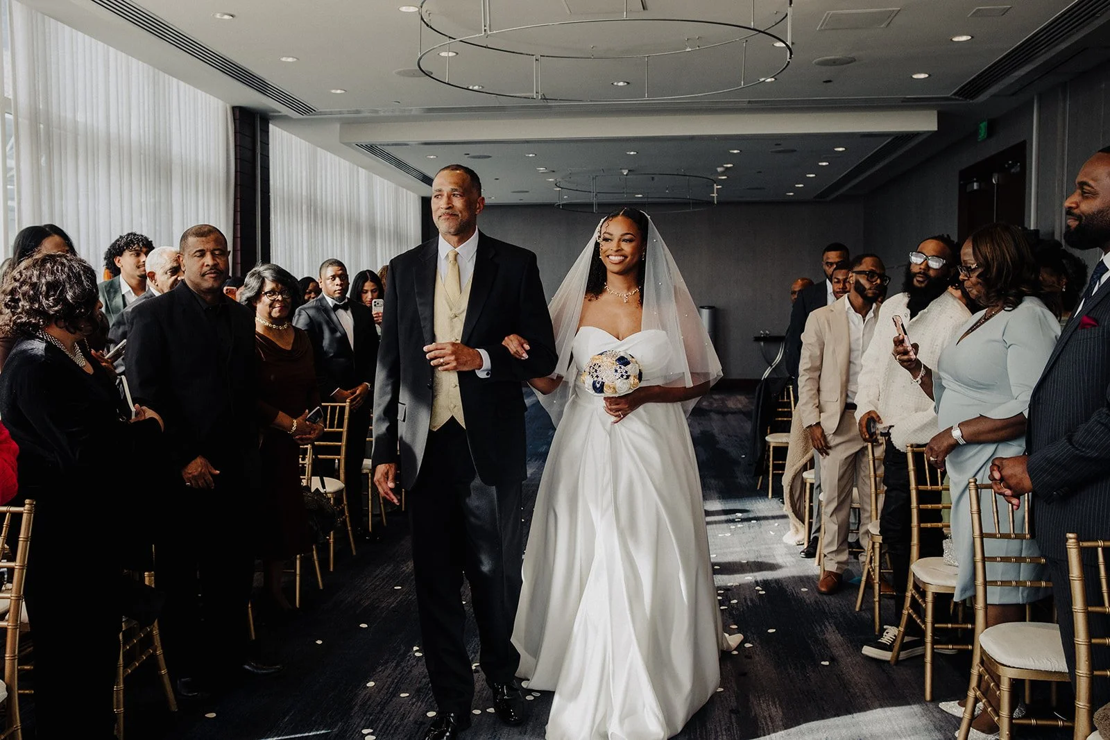 A bride walking down the aisle with her father at a wedding ceremony, surrounded by seated and standing guests in a modern indoor venue.