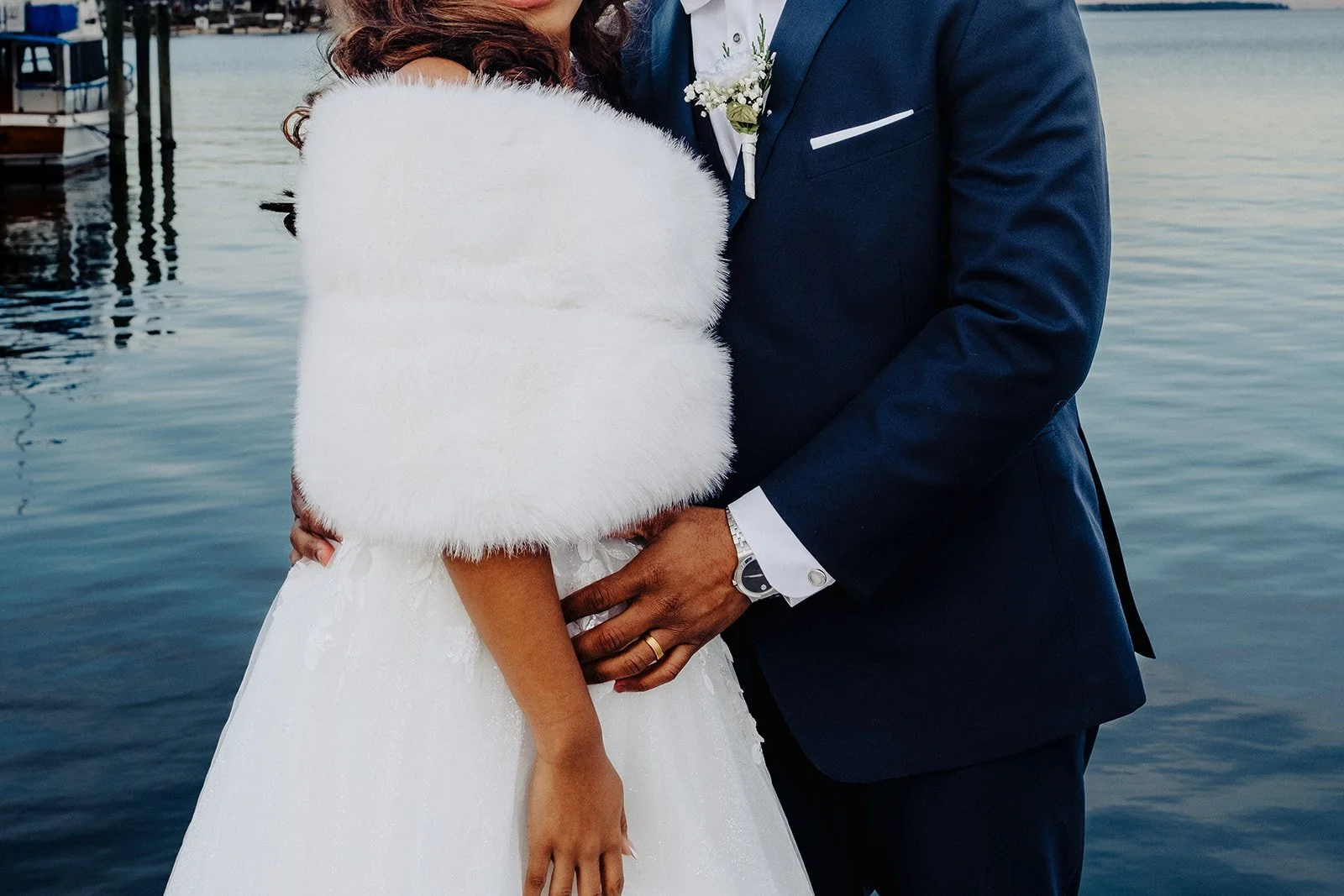 Close-up of a bride and groom holding hands at their wedding by a lake, with the bride wearing a white dress and a fluffy white shawl, and the groom in a navy blue suit with a white shirt and boutonniere.