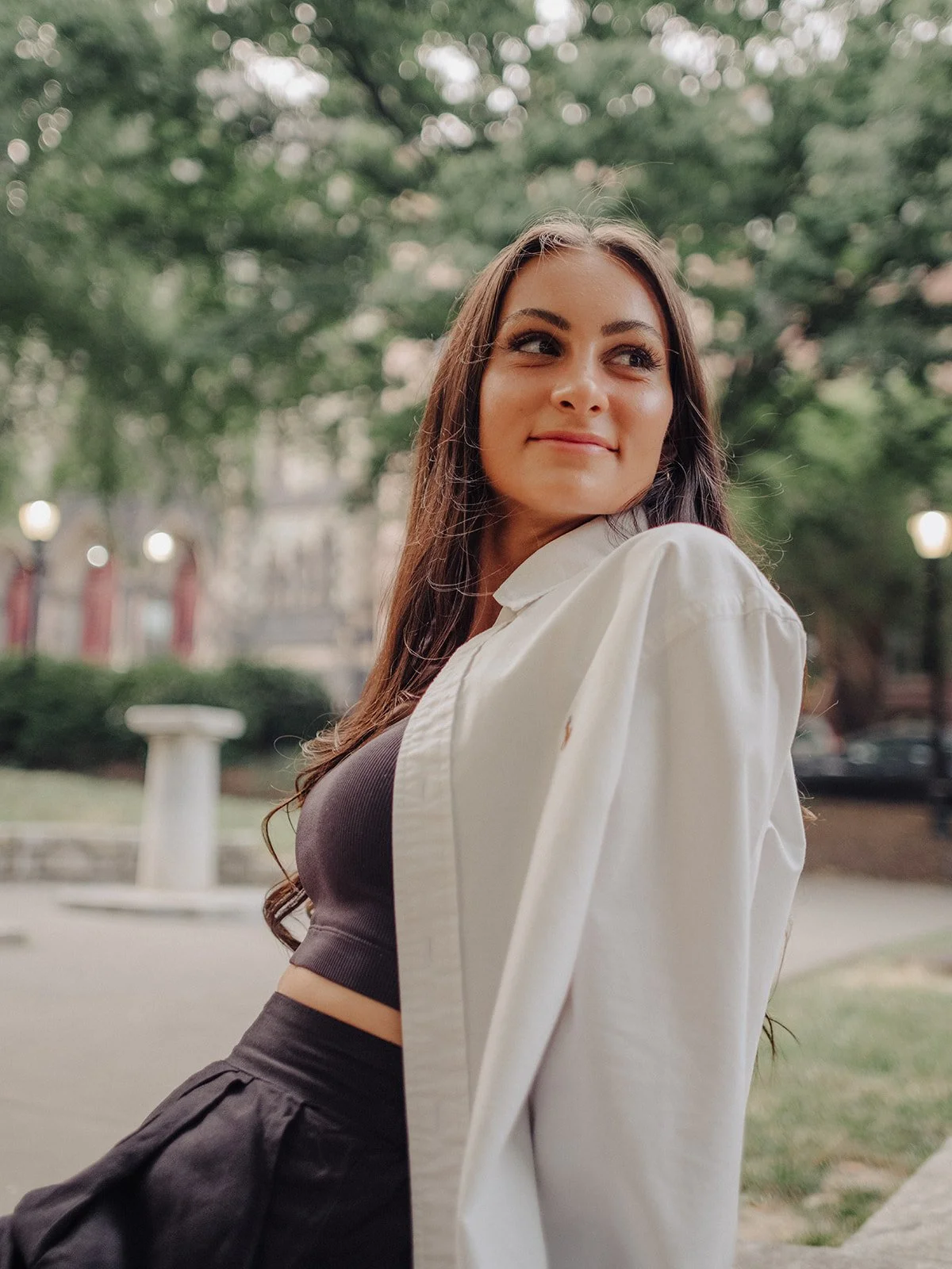 A young woman with long brown hair and makeup smiling softly, looking to her right, standing outdoors in a park with green trees, a white bench, and a building in the background, wearing a black crop top and a white jacket.