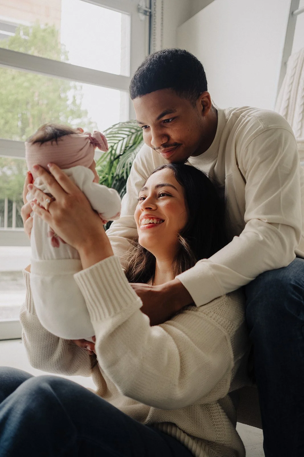 A family, including a woman, man, and a newborn baby, smiling and bonding together indoors near a large window with greenery outside.