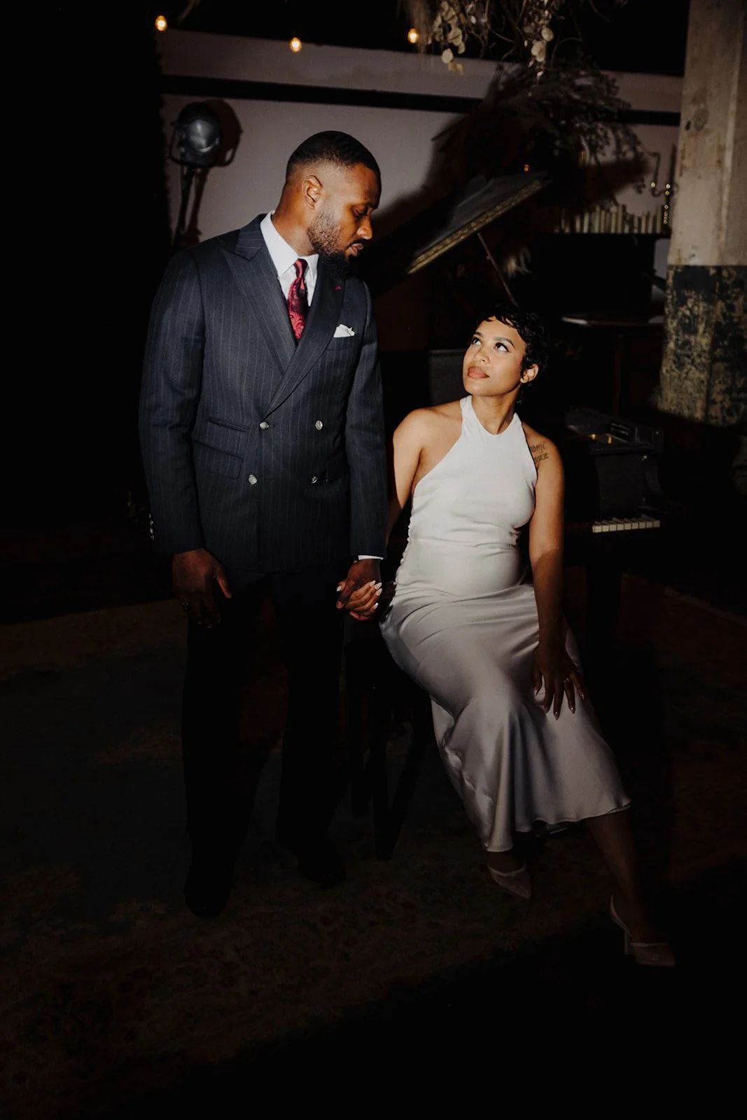 A man in a dark pinstripe suit and a woman in a white dress holding hands in a dimly lit room with a grand piano in the background.