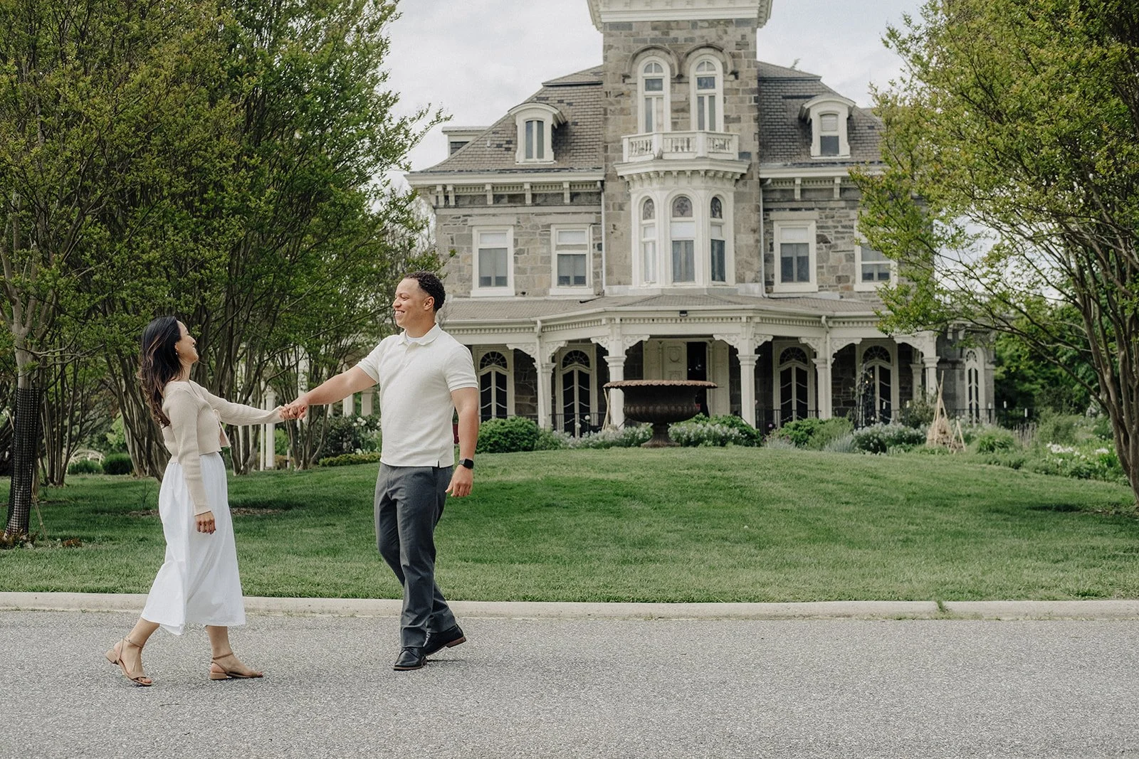 A man and woman holding hands, smiling, dancing on a street in front of a large Victorian-style house with a stone facade, surrounded by green trees and a manicured lawn.