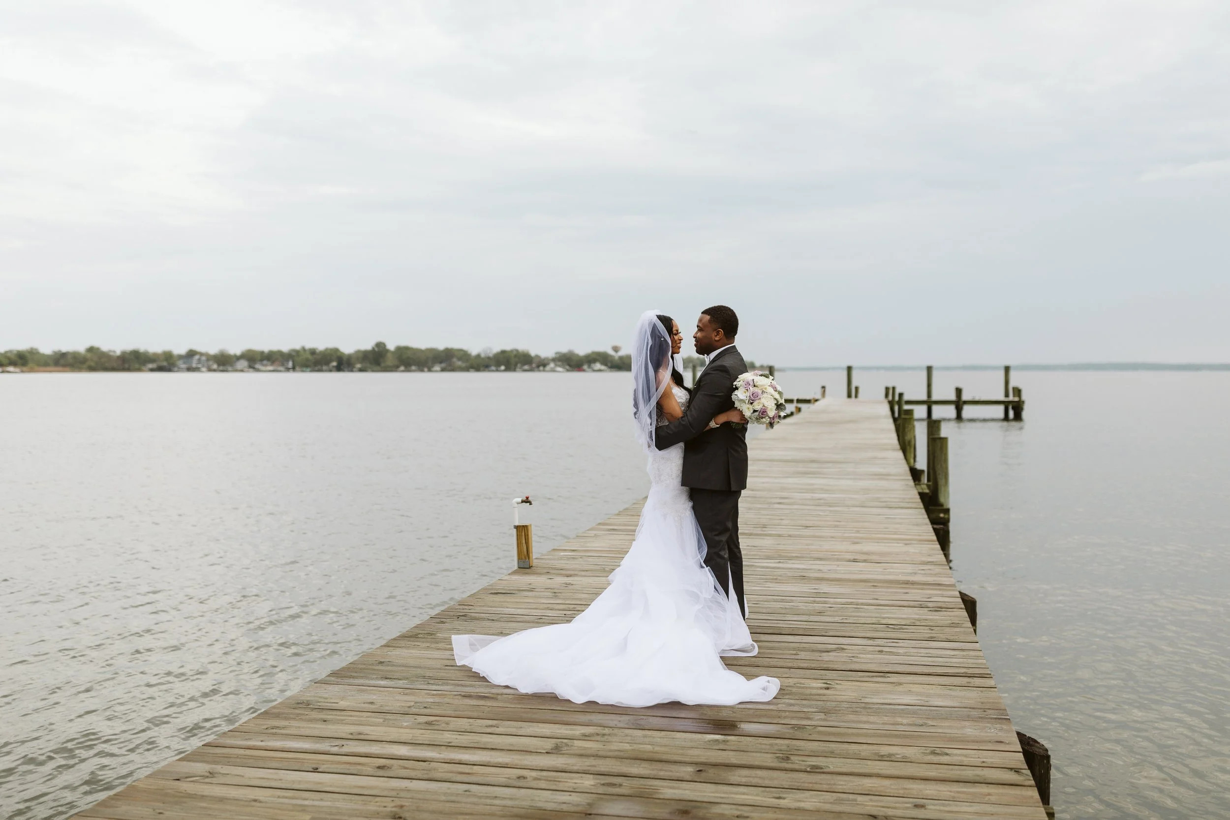 A bride and groom stand on a wooden dock by a lake, gazing at each other, with the bride holding a bouquet of flowers, against a cloudy sky background.