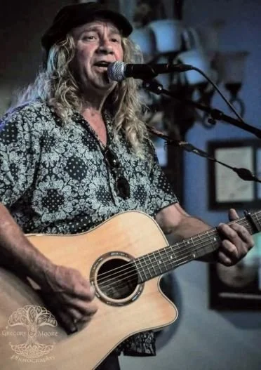 An older woman with long curly gray hair singing into a microphone while playing an acoustic guitar in a dimly lit setting, possibly a small performance venue or bar.
