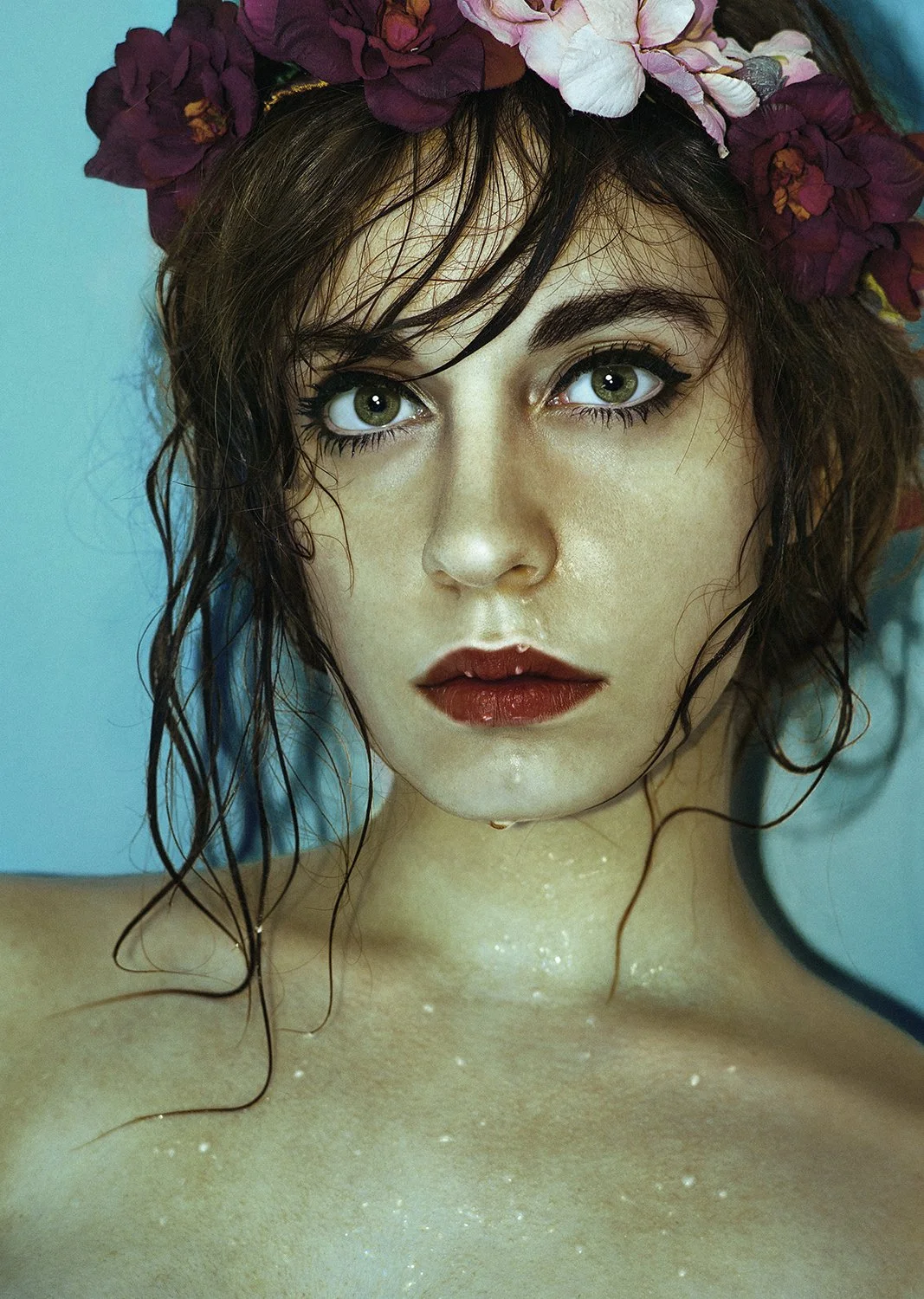 A close-up photo of a woman with wet hair, green eyes, and dark makeup, wearing a flower crown made of purple and pink flowers, against a blue background.