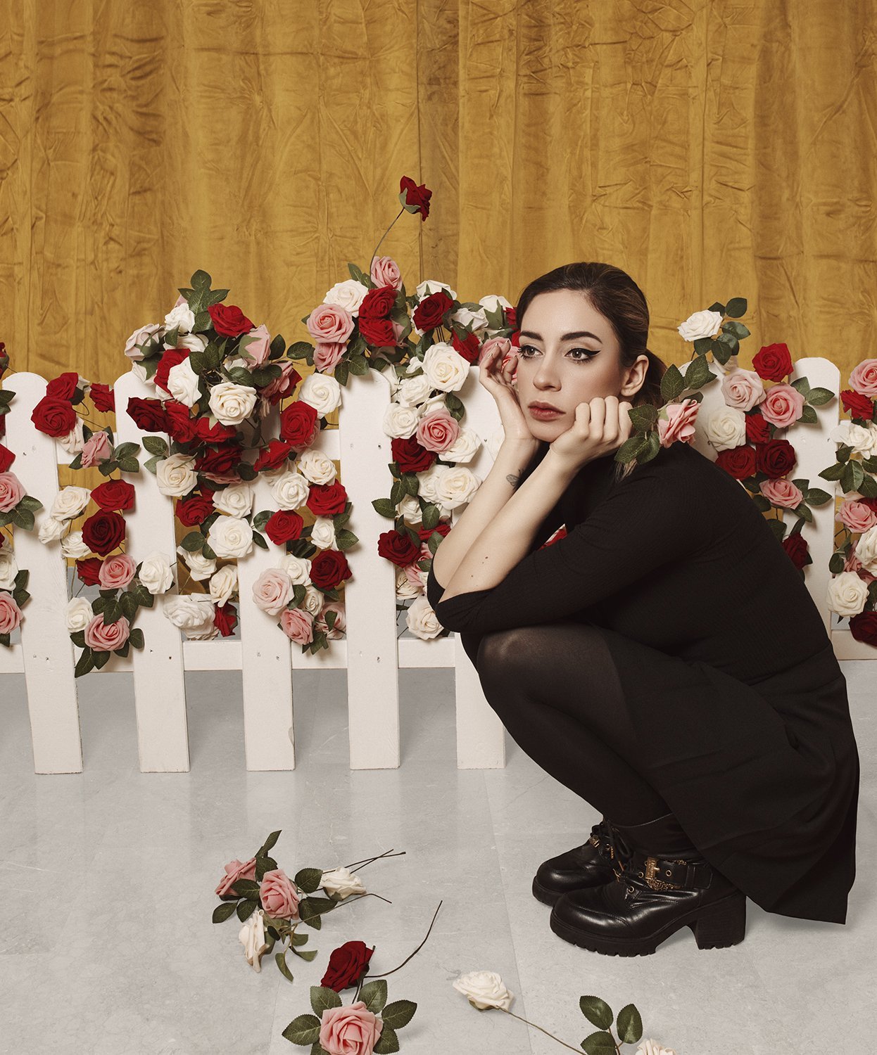 A woman with dark hair and winged eyeliner squats in front of a white picket fence covered with red, pink, and white roses, with some roses on the ground, against a golden curtain background.