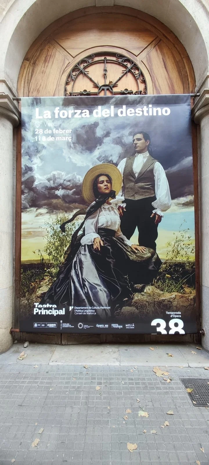 A poster advertising an opera titled 'La fuerza del destino', showing a man and a woman dressed in historical clothing, set outdoors with a cloudy sky background. The woman is sitting with a large sunhat and black dress, the man is standing behind he