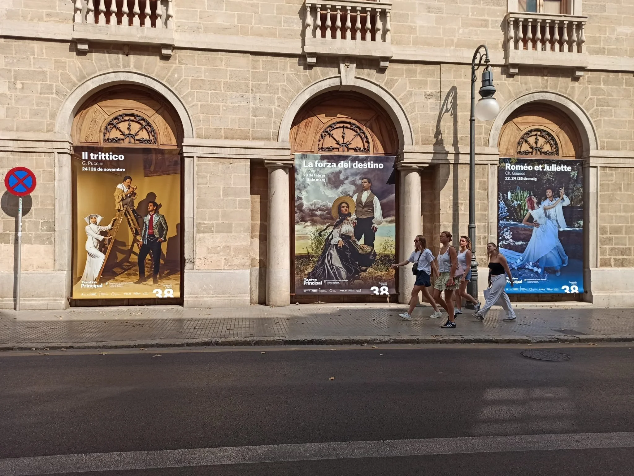 Street view showing three large posters advertising operas: 'Il trittico' by G. Puccini, 'La forza del destino', and 'Roméo et Juliette', with a group of five women walking past in daylight, a streetlamp, and a no-parking sign on the sidewalk.