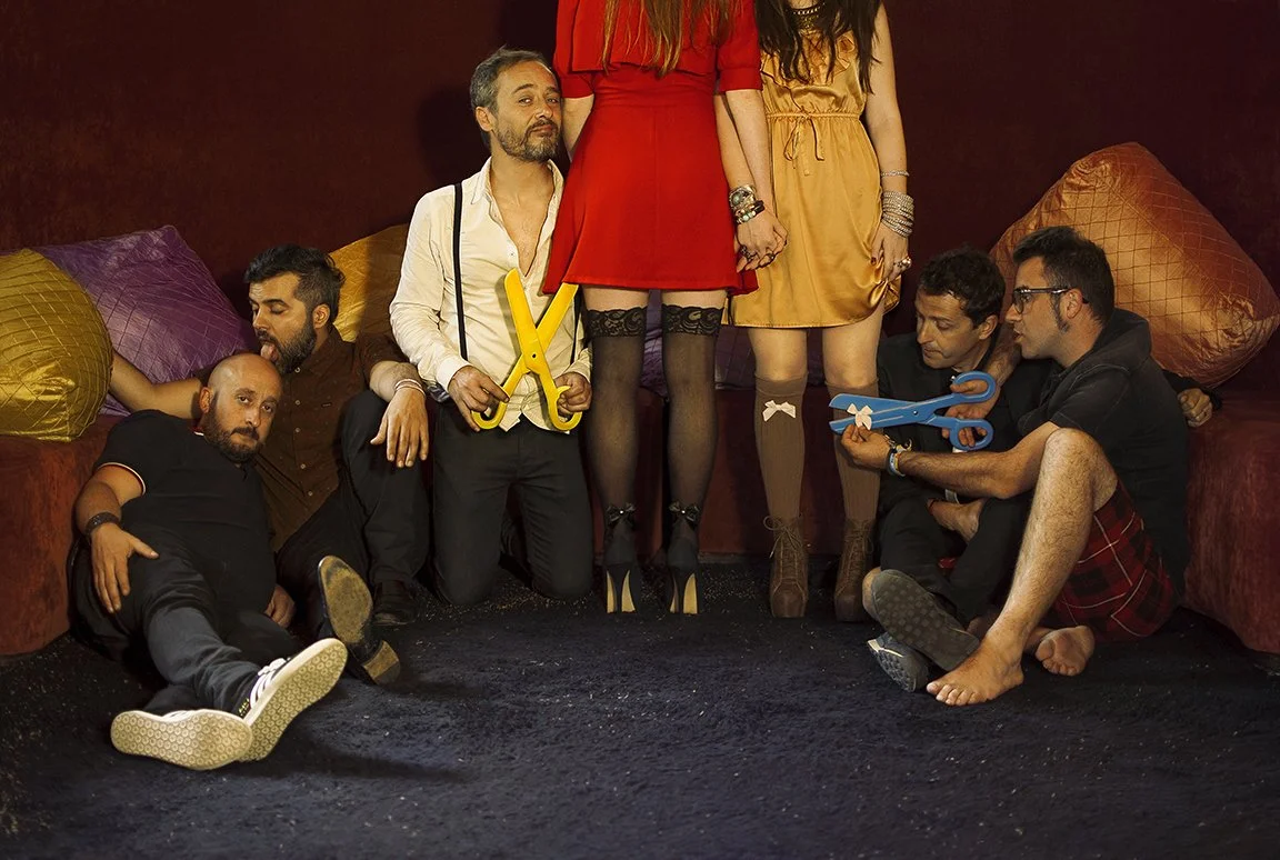 Group of seven people sitting and standing on a dark carpeted floor against a dark red wall, with large pillows behind them. The men are dressed casually, some holding toy scissors, and two women stand in the middle wearing dresses and stockings.