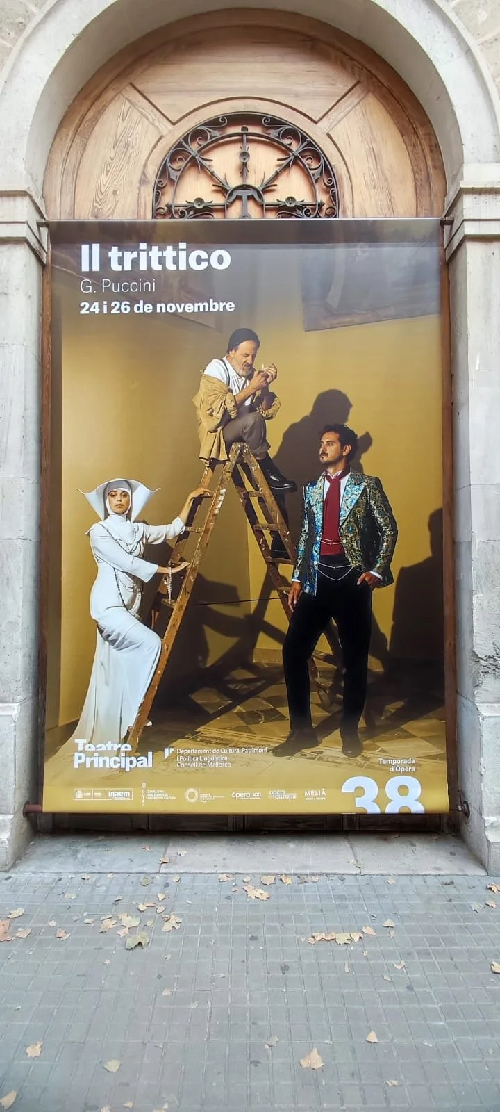 Poster advertising Puccini's opera "Il Trittico" showing three characters in theatrical costumes, with one sitting on a ladder, one standing with hand on hip, and a woman in a white costume holding the ladder.
