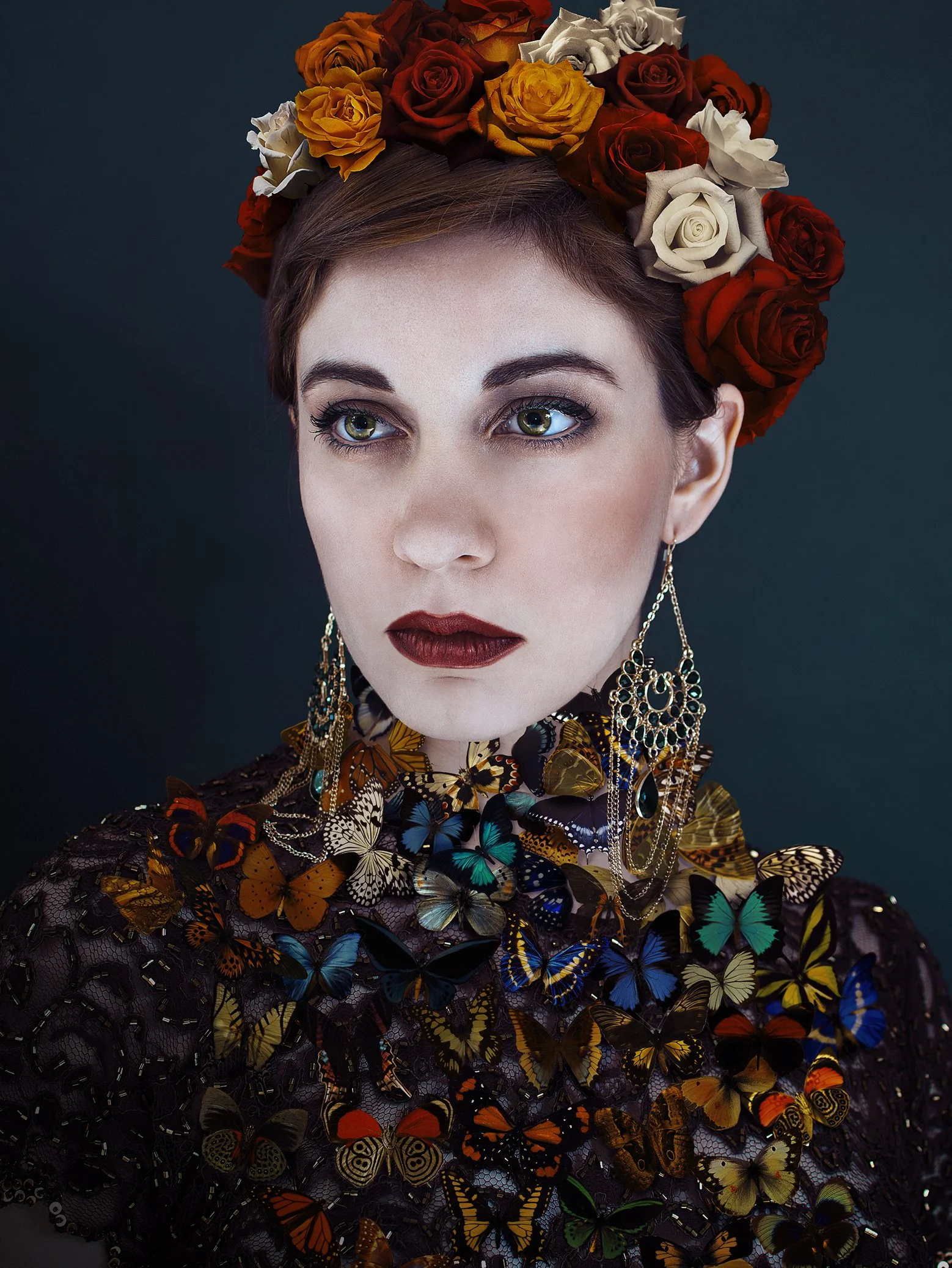 A woman with short reddish-brown hair wears a crown of red, orange, white, and beige roses, large ornate earrings, and a dress adorned with colorful butterflies.