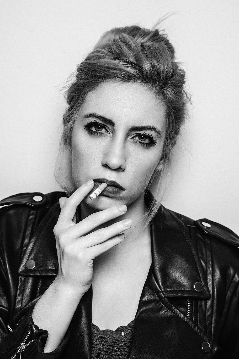 Black and white portrait of a woman with intense expression, smoking a cigarette, wearing a leather jacket, with hair styled in an updo.