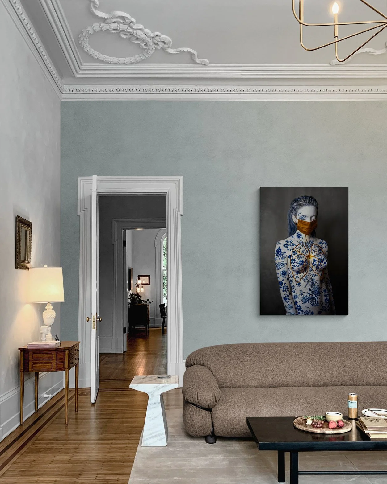 Elegant living room with ornate ceiling design, a modern chandelier, beige sofa, white marble side table, and artwork featuring a person with floral patterns on the wall. Open doorway leads to a hallway with wooden flooring.
