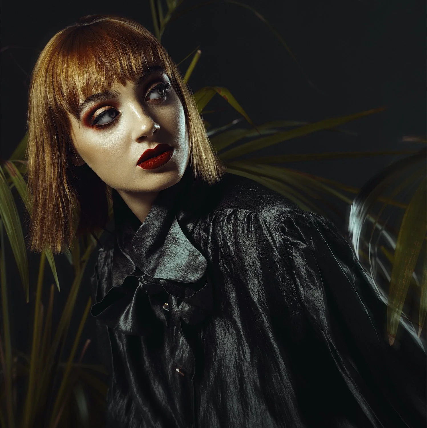 A woman with short brown hair and bold makeup, wearing a shiny black blouse, sits among dark green foliage in a dark setting.