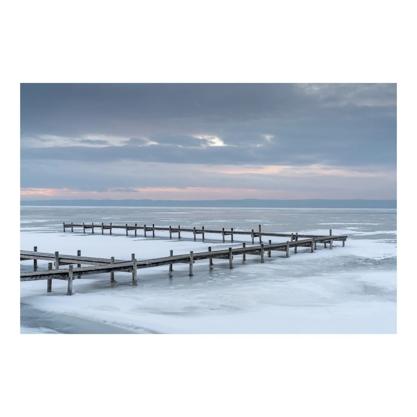 When ice claims the shallow waters of Neusiedler See, the summer boardwalks become pathways through a monochrome dreamscape. Here at Podobeach, soft dusk light illuminates the intersection of structure and nature, where human design meets the raw bea