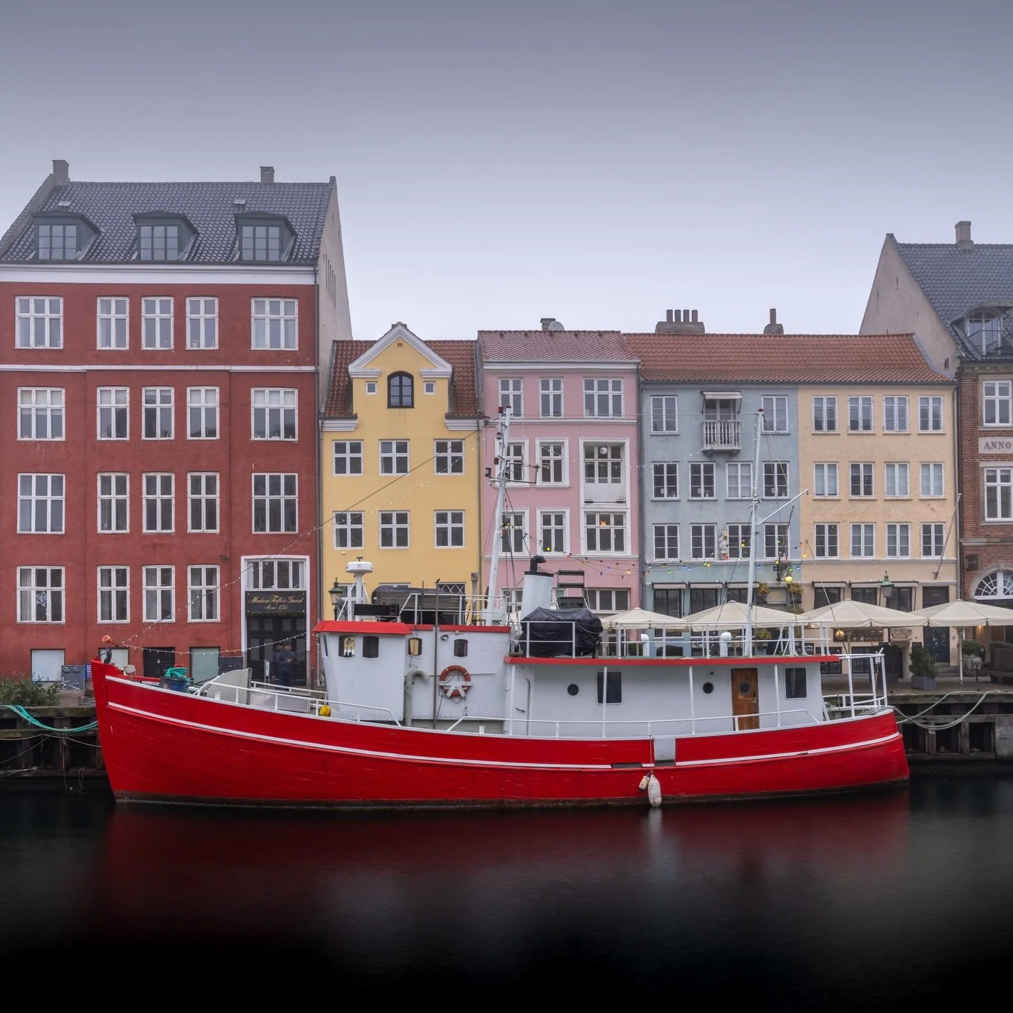 Stillness settles over the water while stories from centuries past linger in every weathered fa&ccedil;ade. Nyhavn&rsquo;s iconic harbor transforms under muted skies, where a single vessel holds the frame against townhouses that once sheltered sailor