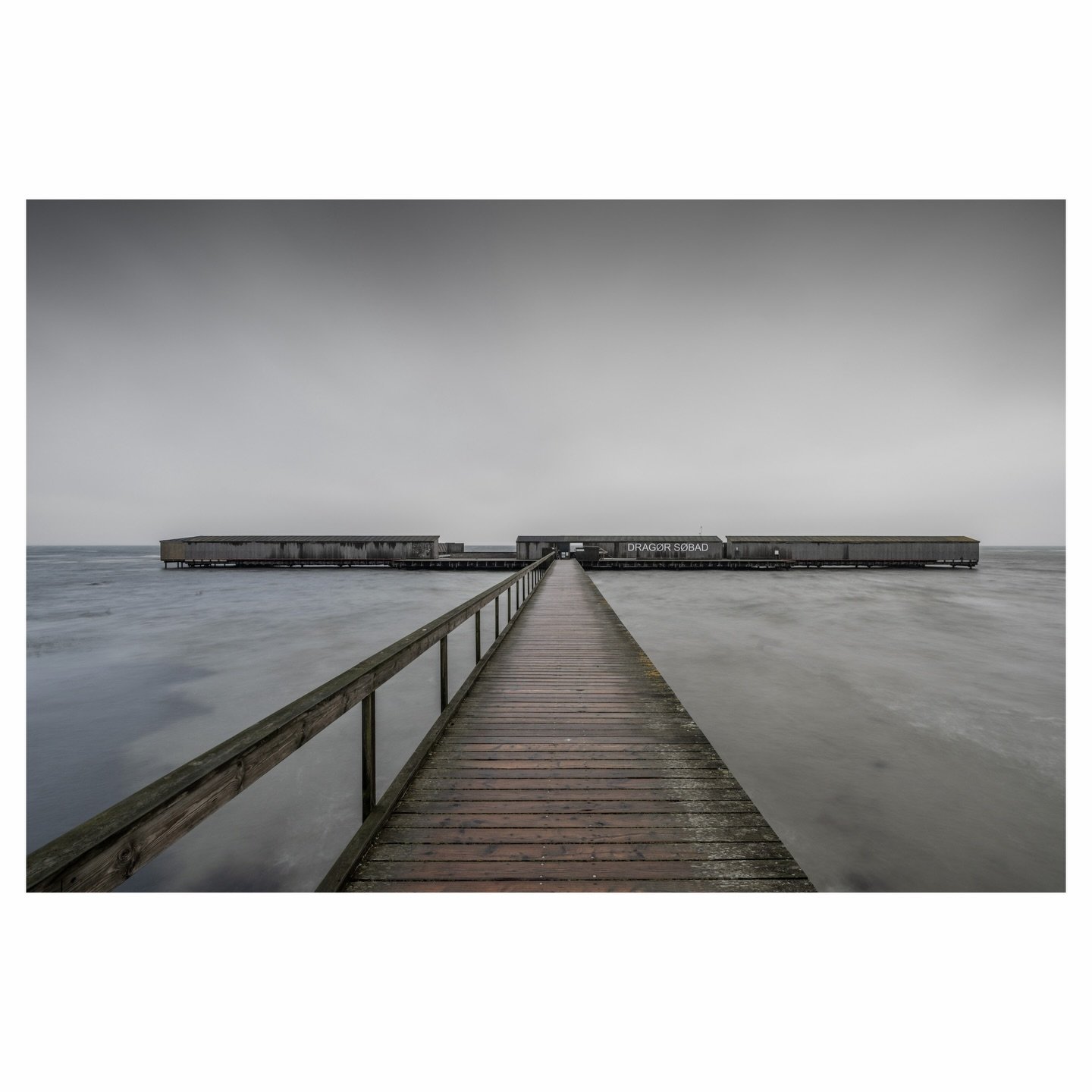 The first light of a Sunday morning reveals the quiet geometry of Drag&oslash;r S&oslash;bad, where separated walkways lead swimmers toward enclosed basins and twin saunas hidden within those wooden walls. Long exposure transforms the &Oslash;resund 