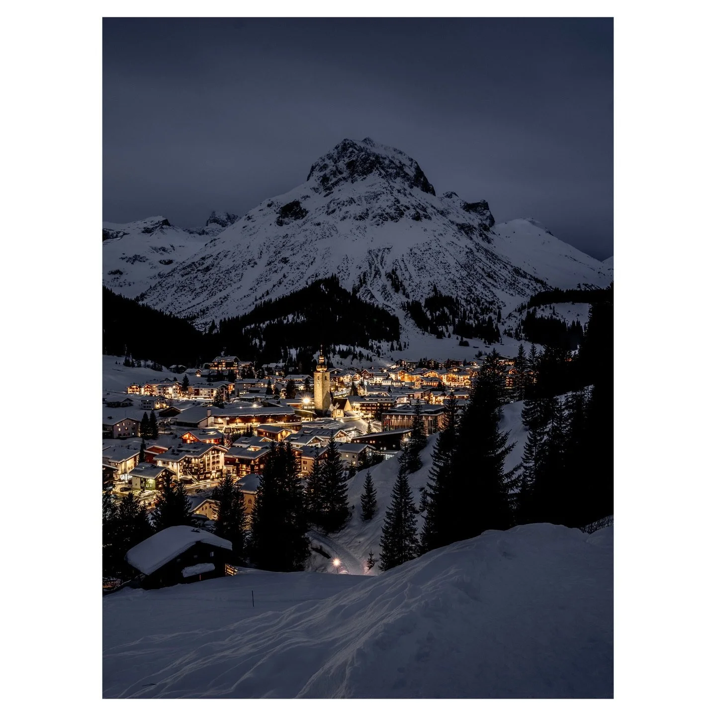 Lech am Arlberg after sunset, when the slopes go quiet and the village center takes over. Viewed from above, a few hundred lights glow against the Omeshorn as the valley settles into its calmest hour. No sound, no rush, just snow and warmth in perfec
