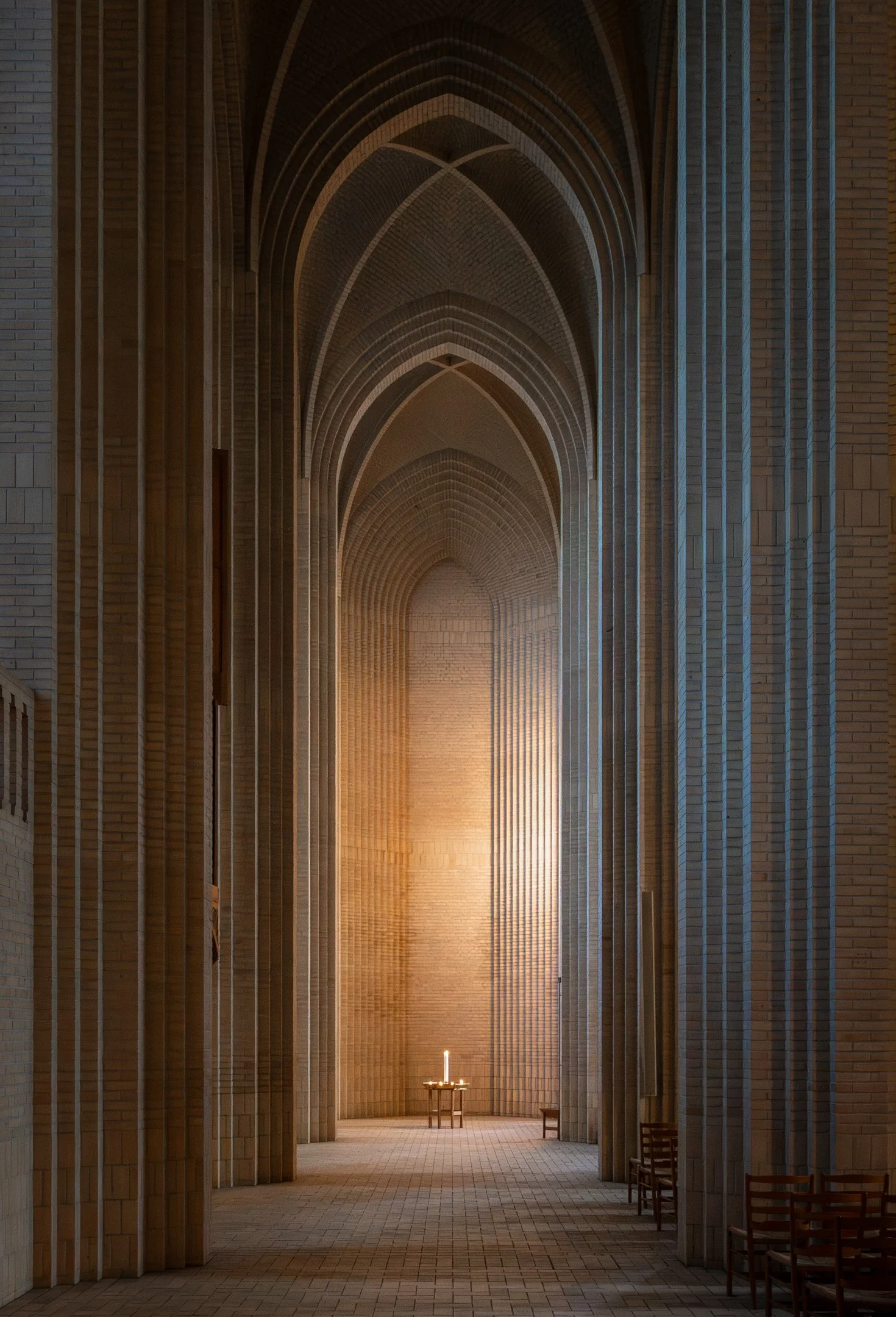 Interior view of a church or cathedral with high vaulted ceilings and brick walls, illuminated by a candle on a stand at the end of a narrow aisle.