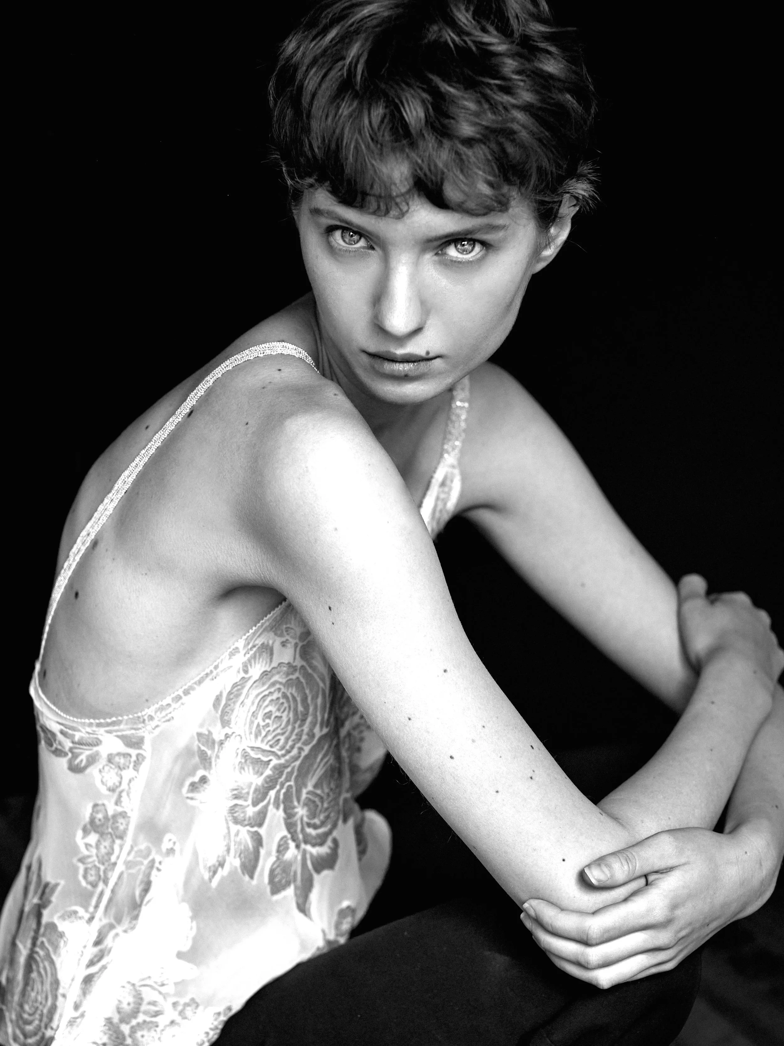 A black and white photograph of a young woman with short, wavy hair, looking directly at the camera. She is wearing a lace camisole and has a serious expression. Her arms are crossed and resting on her knees, with her hands clasped.