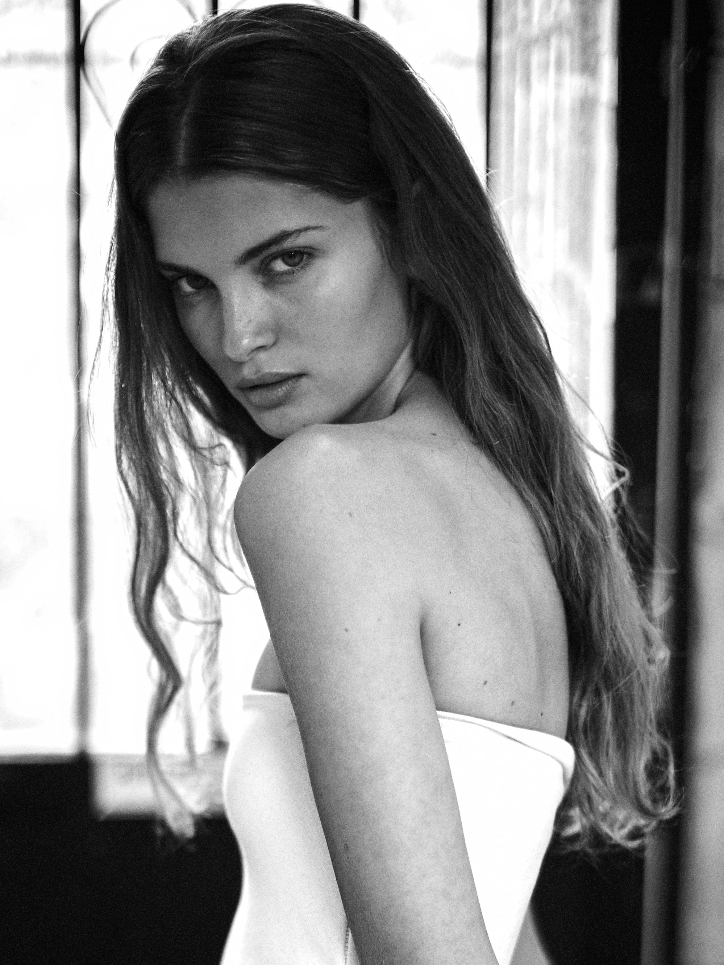 A black-and-white photograph of a young woman with long hair, looking over her shoulder at the camera, in front of a window with light coming through.