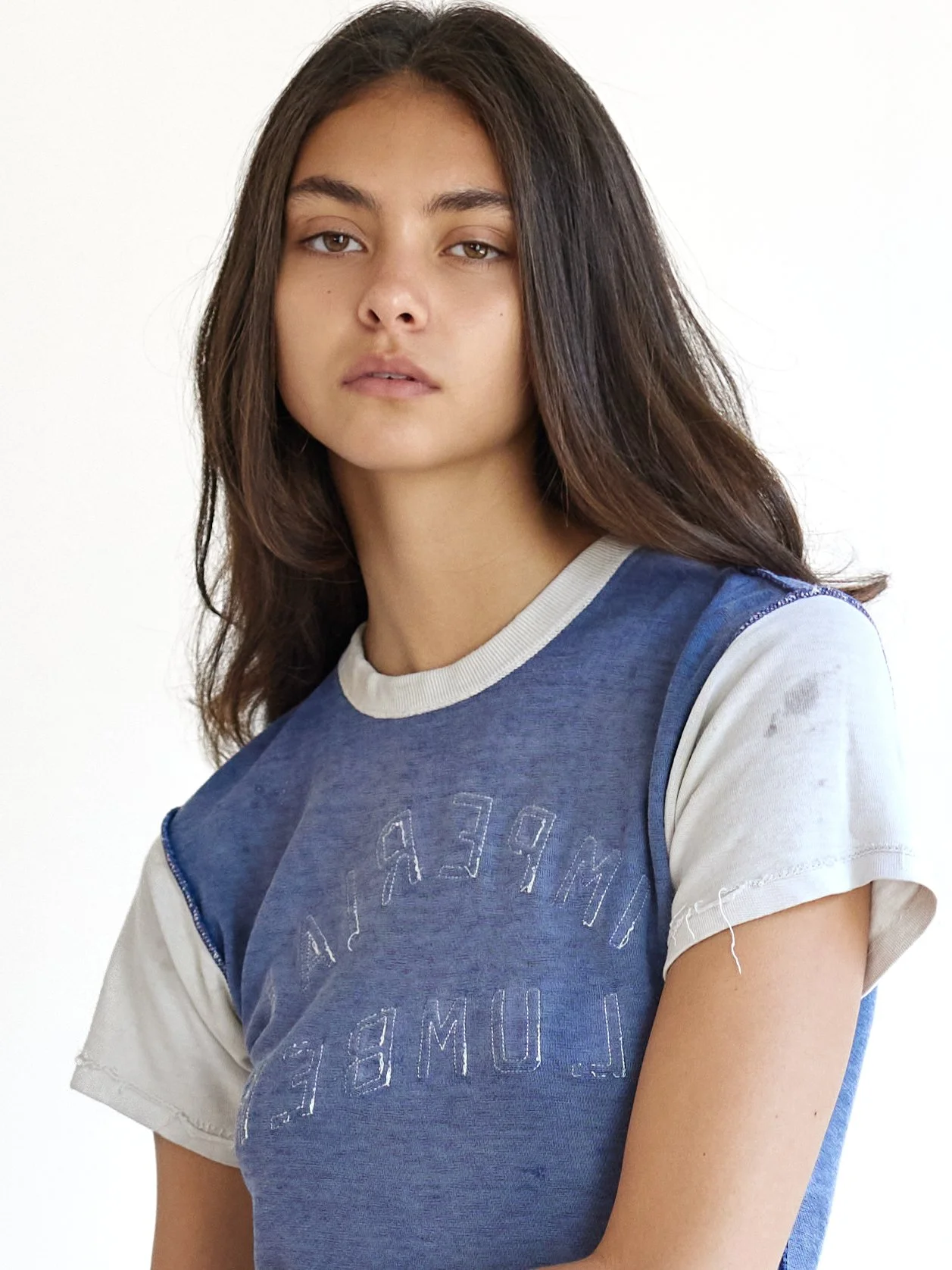 Portrait of a young woman with shoulder-length brown hair wearing a blue and white t-shirt standing against a plain white background.