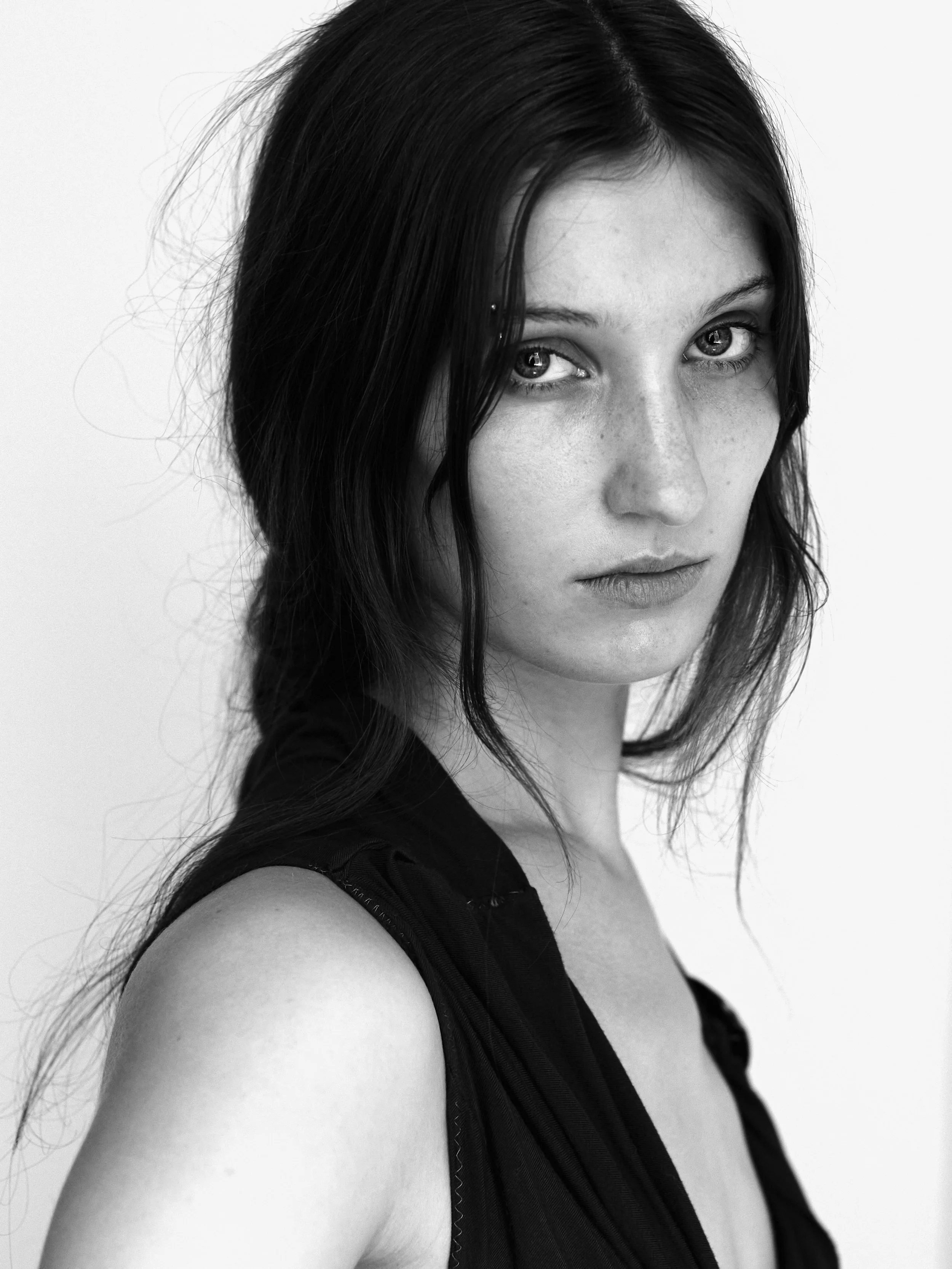 A black and white portrait of a woman with long dark hair, wearing a sleeveless top, looking directly at the camera with a serious expression.