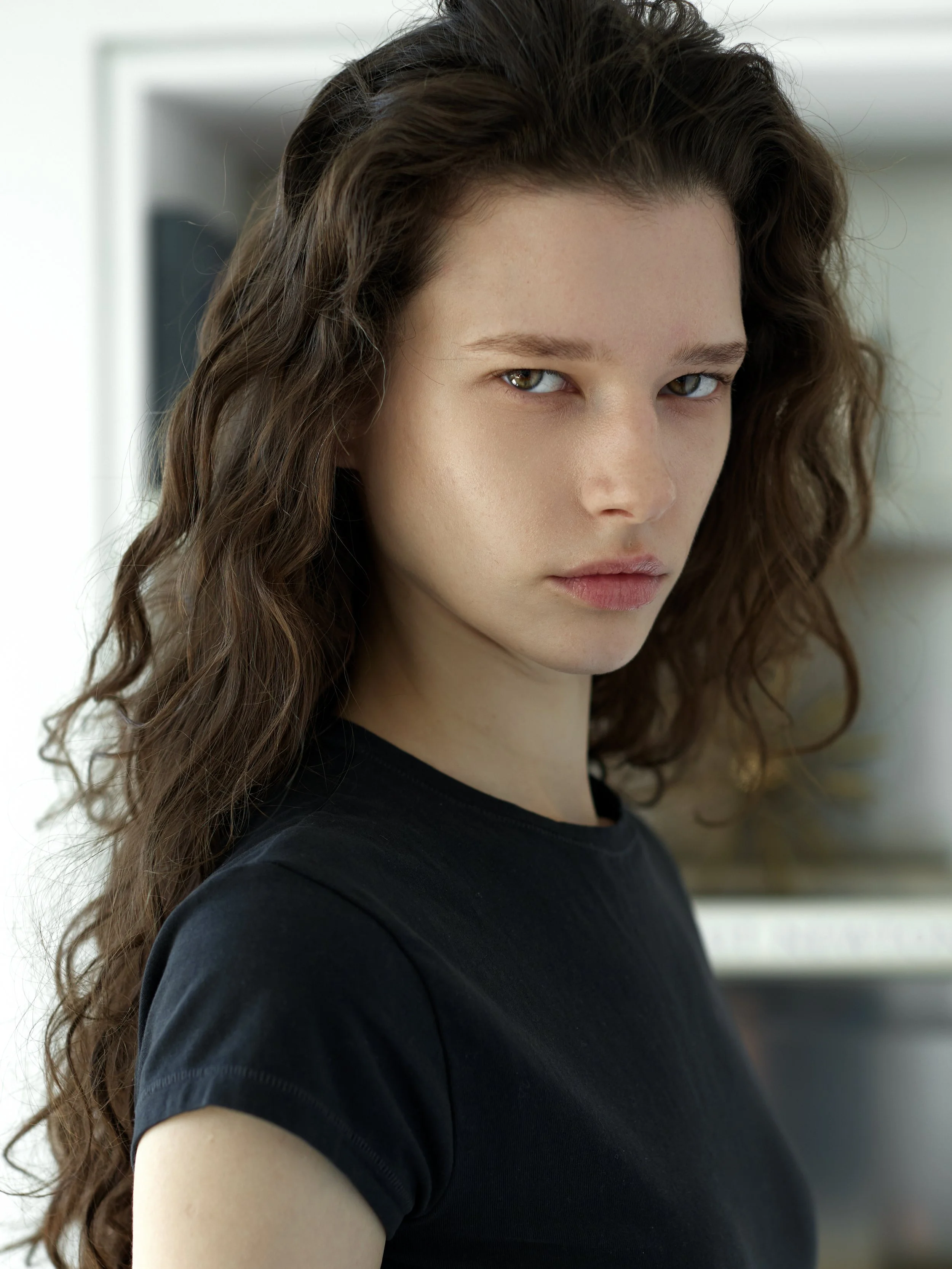 Close-up of a young woman with wavy brown hair and light skin, wearing a black shirt, looking at the camera with a neutral expression.